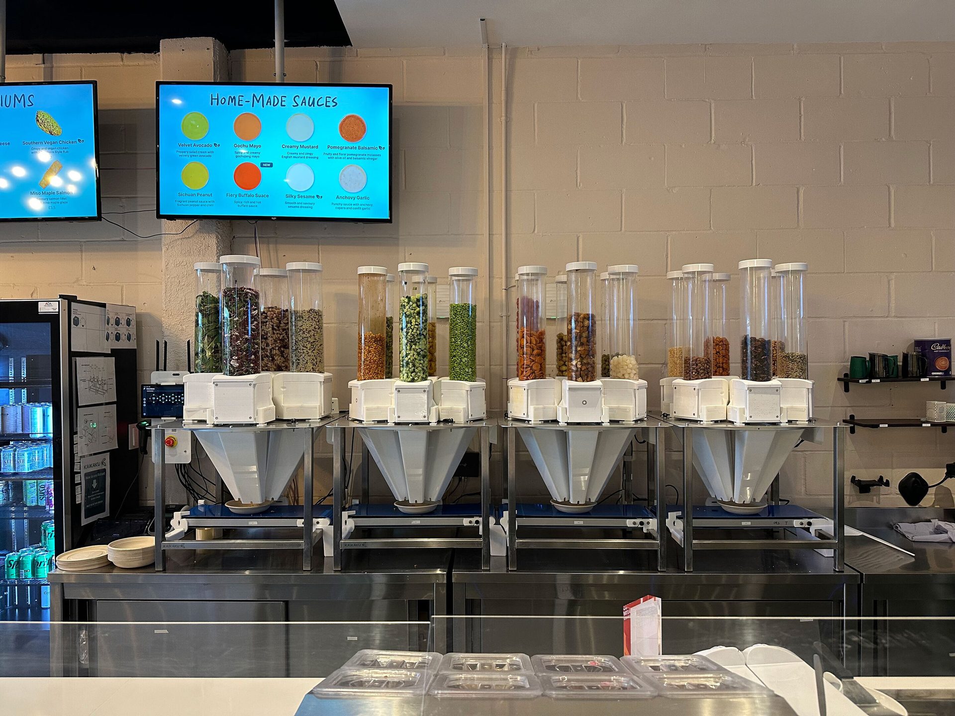 A modern counter with KAIKAKU ingredient dispensers filled with nuts and grains, alongside digital menu screens.