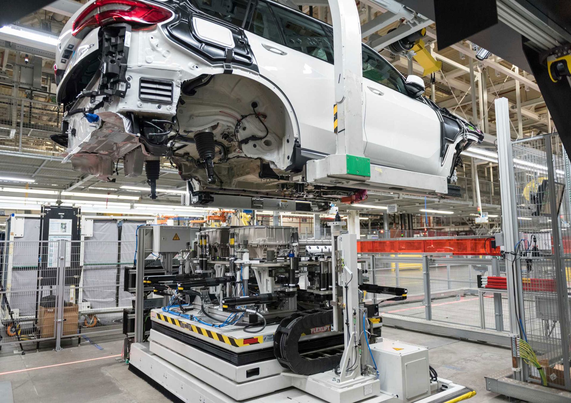 Partially assembled white SUV body suspended above an automated assembly machine in a factory.