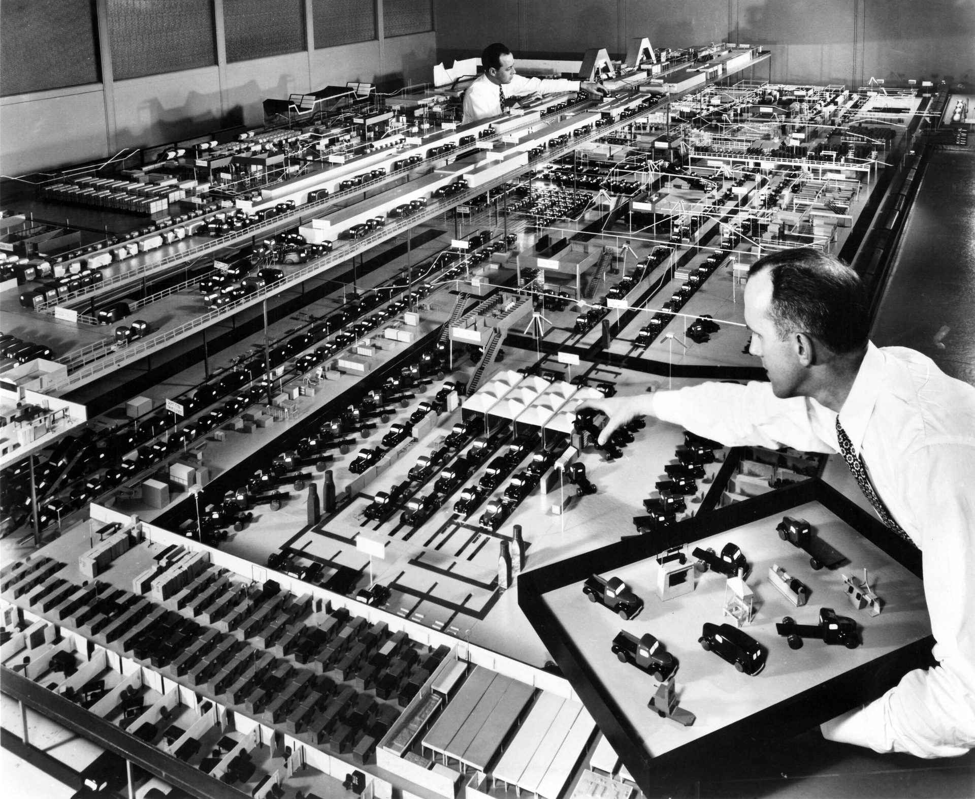 Two men arranging miniature cars on a massive, detailed model of a city or factory layout.