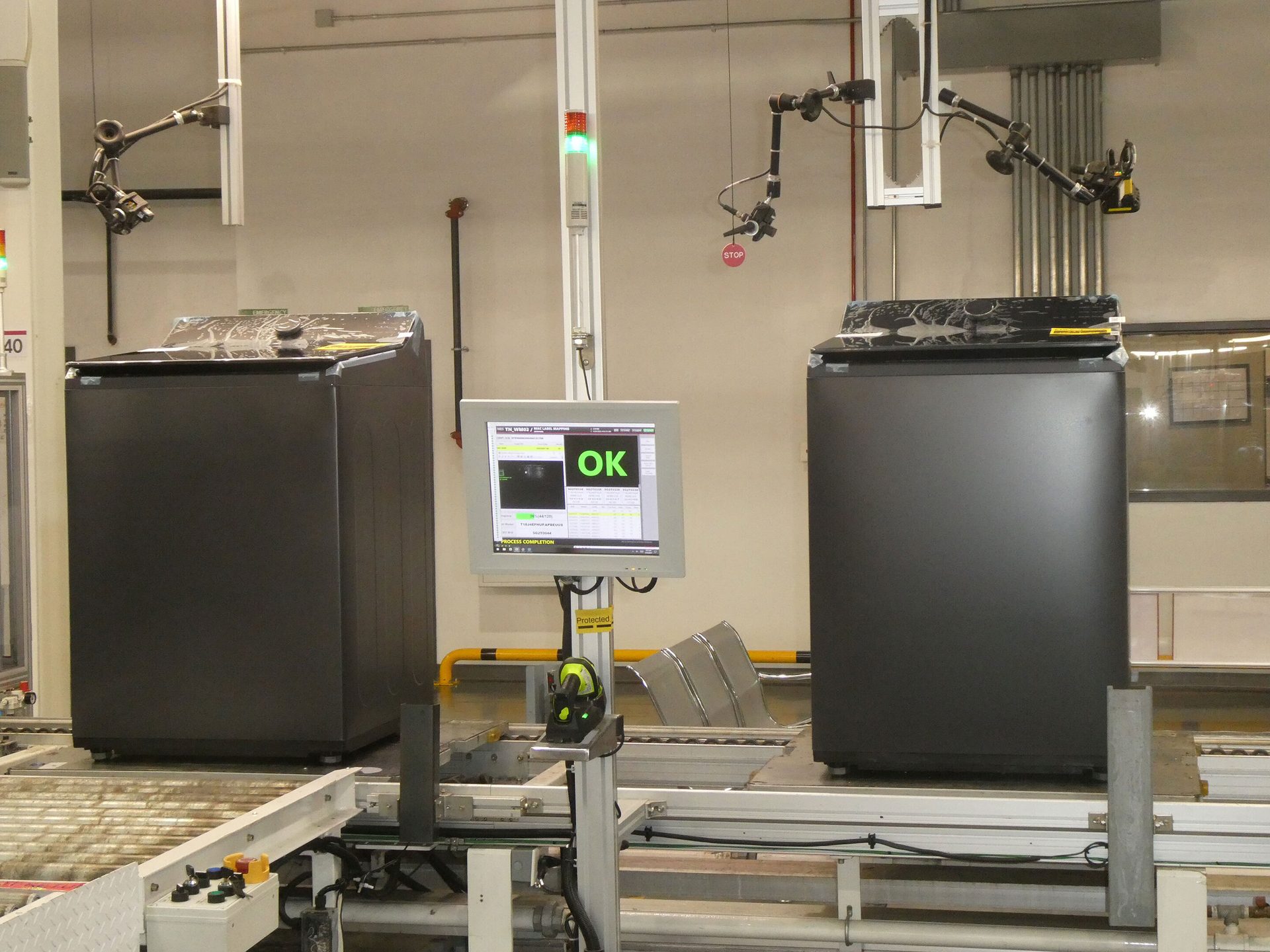 Industrial scene with two large dark machines on conveyor belts, robotic arms, and a monitor displaying "OK".