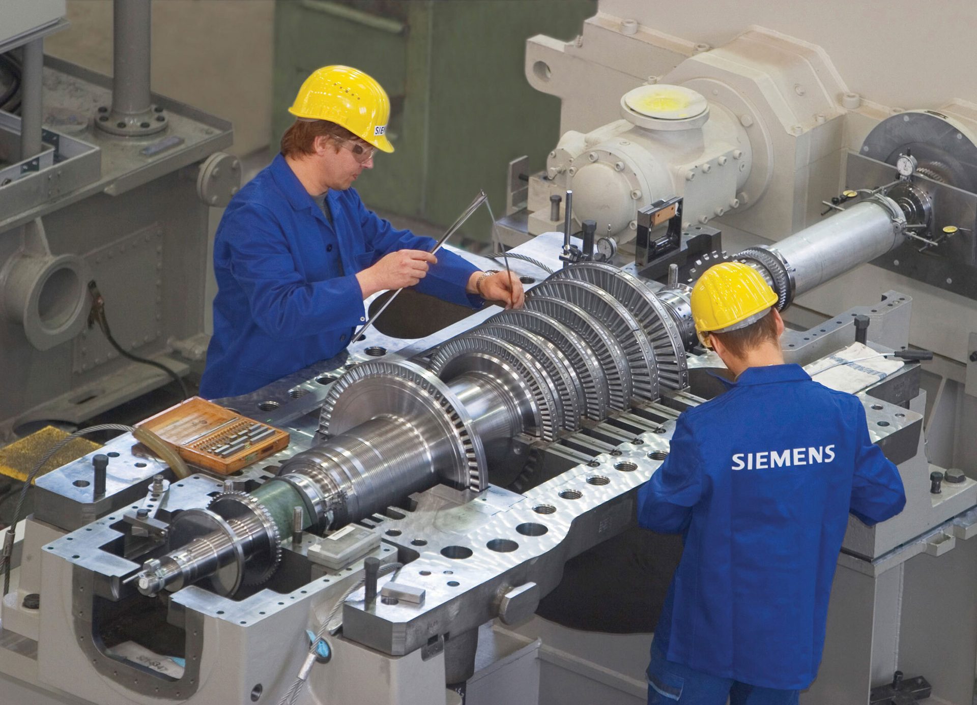 Two Siemens workers in hard hats inspect and assemble a large turbine rotor.