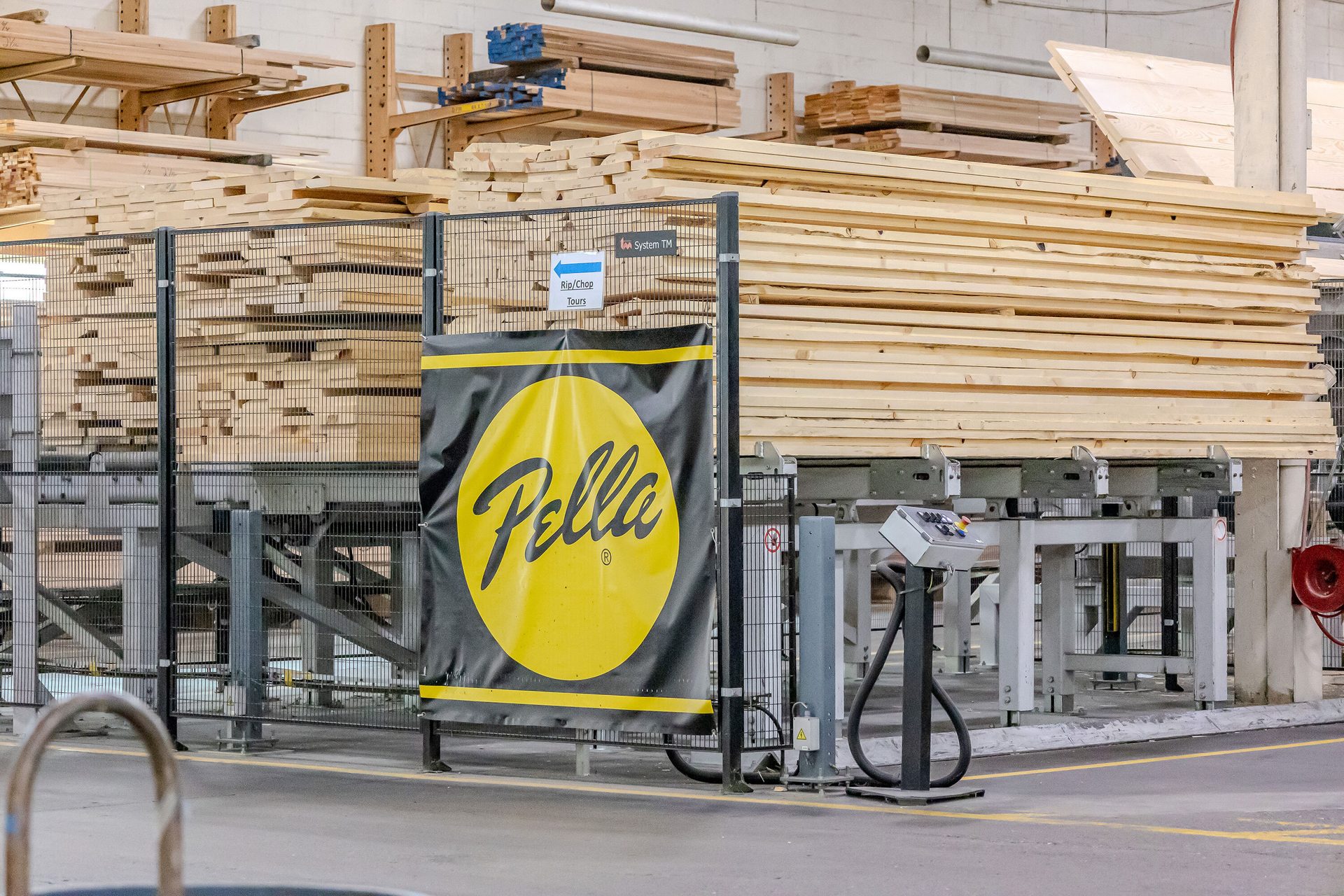Stacks of wood planks on industrial machinery, with a large Pella logo banner prominently displayed.
