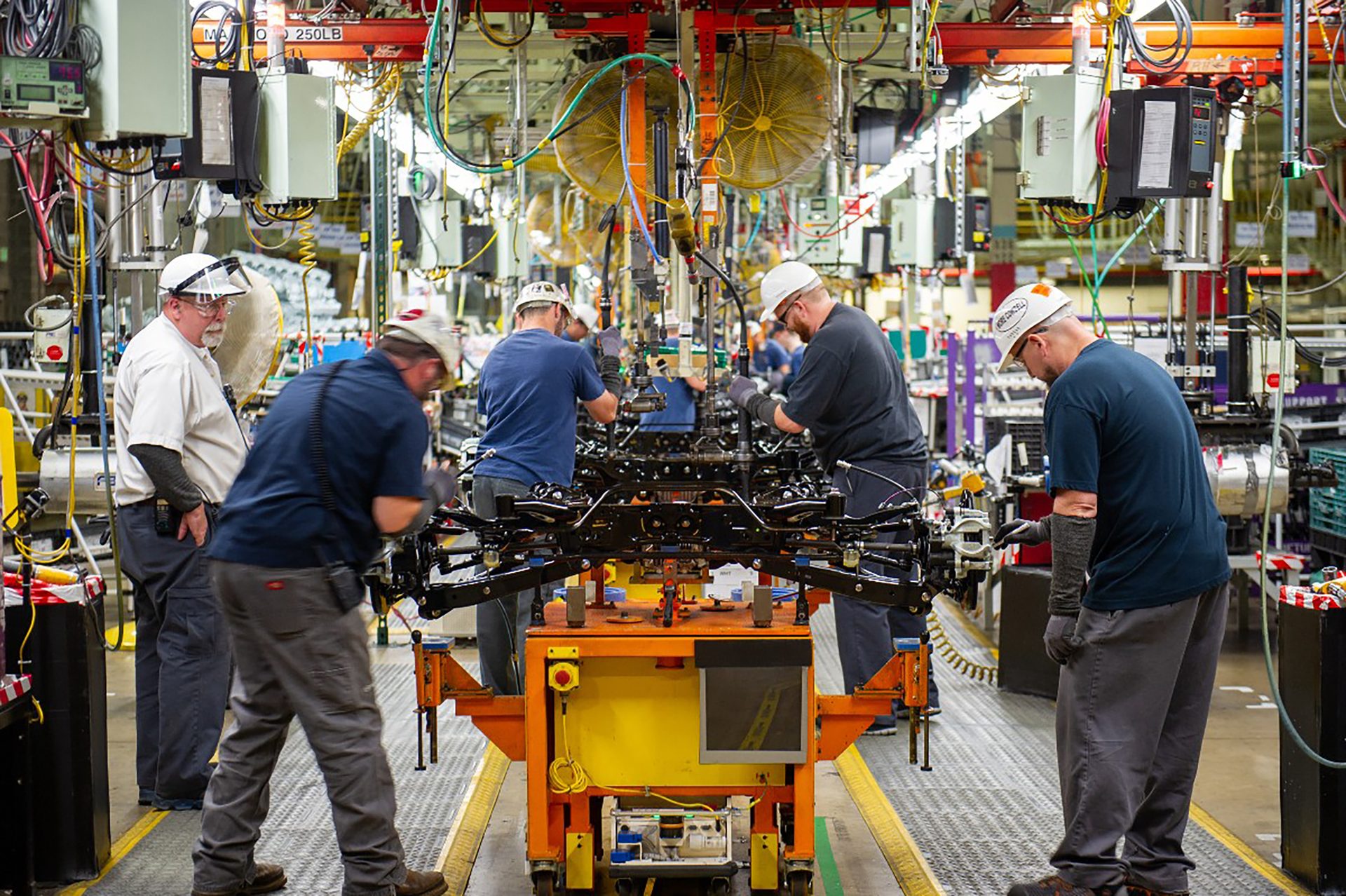 Five workers assemble vehicle parts on a cart in a busy factory.