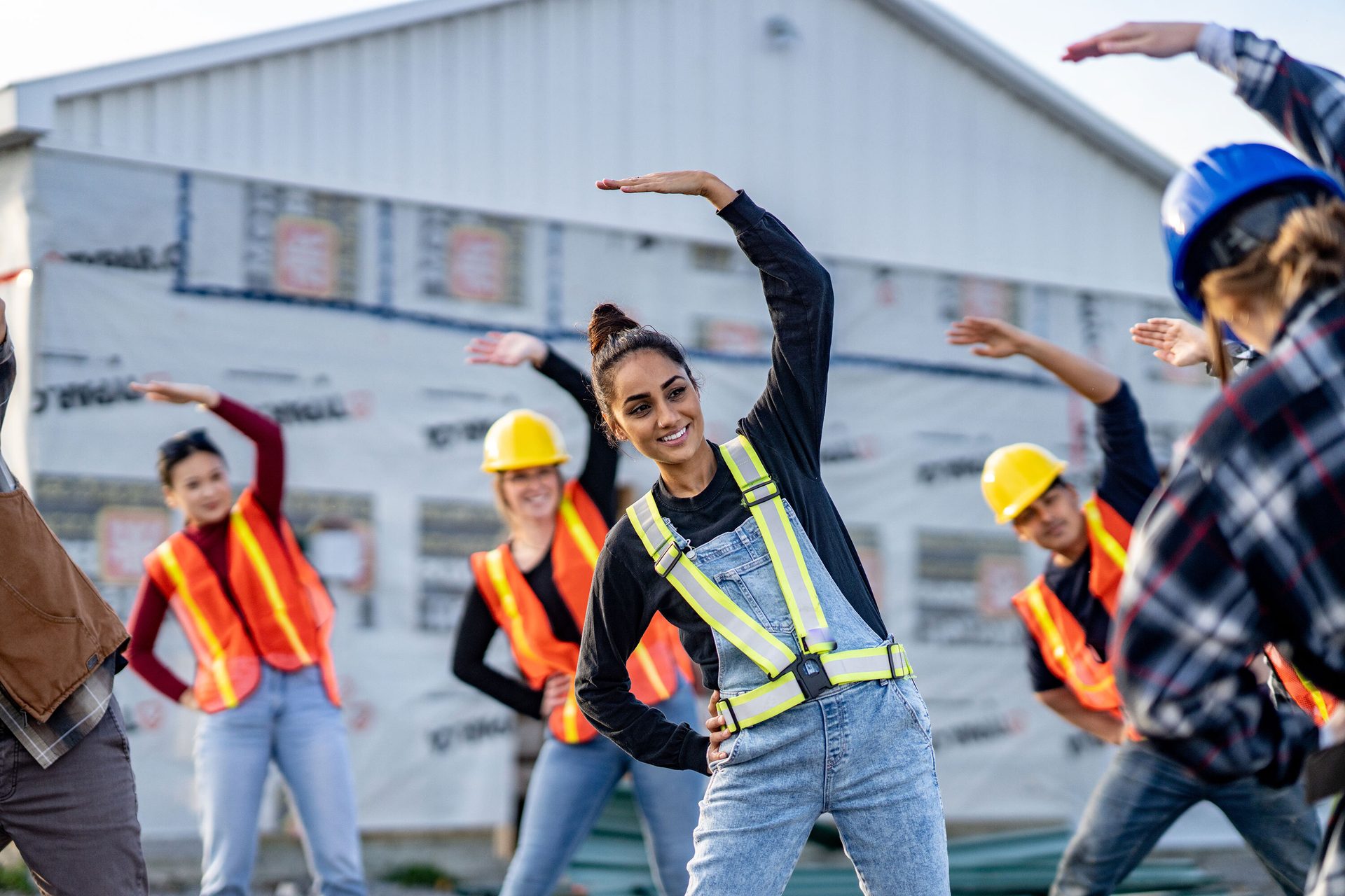 Construction workers in safety gear stretch at a construction site.