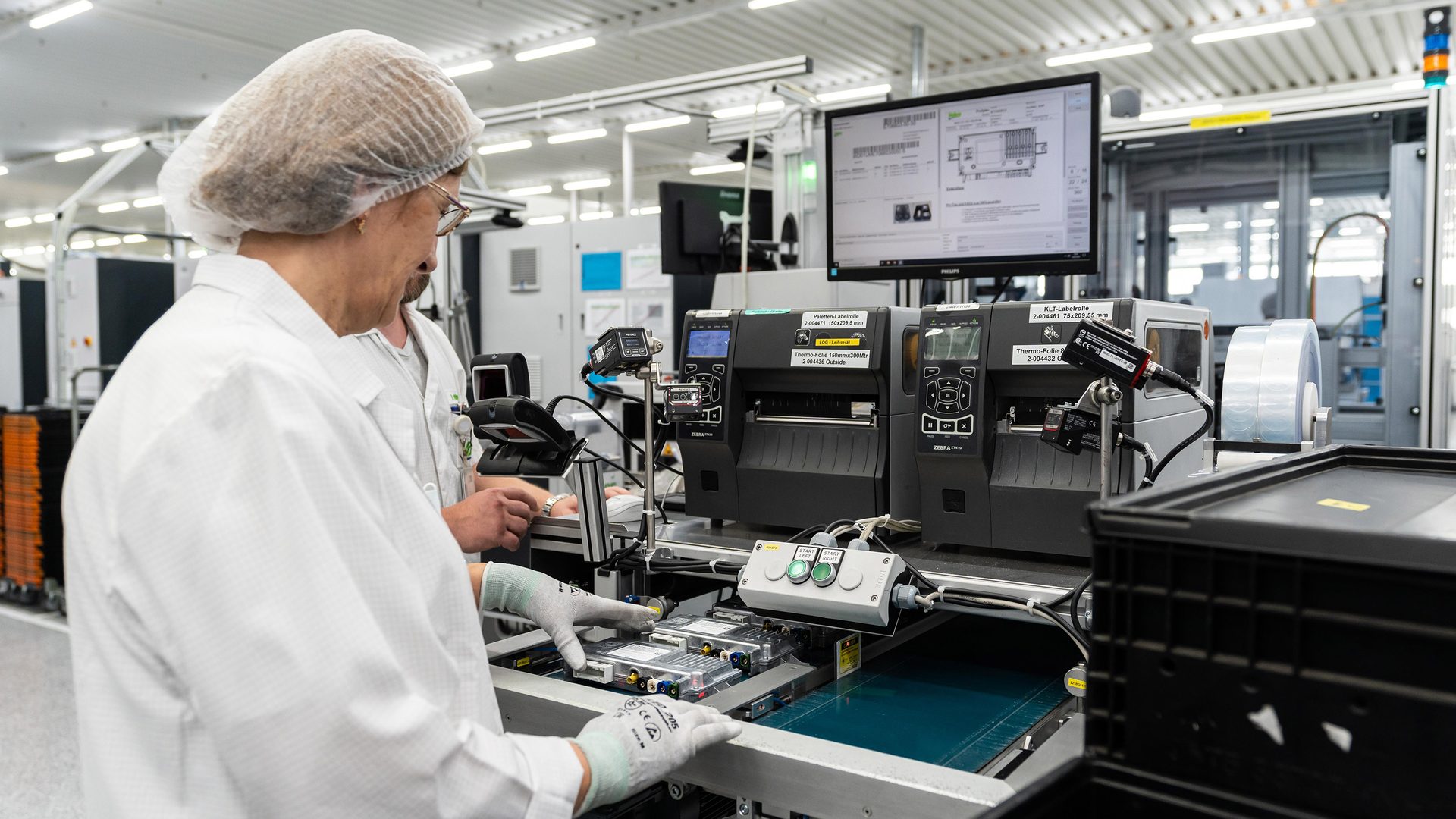 Person in cleanroom attire assembling electronics on an automated line with label printers.