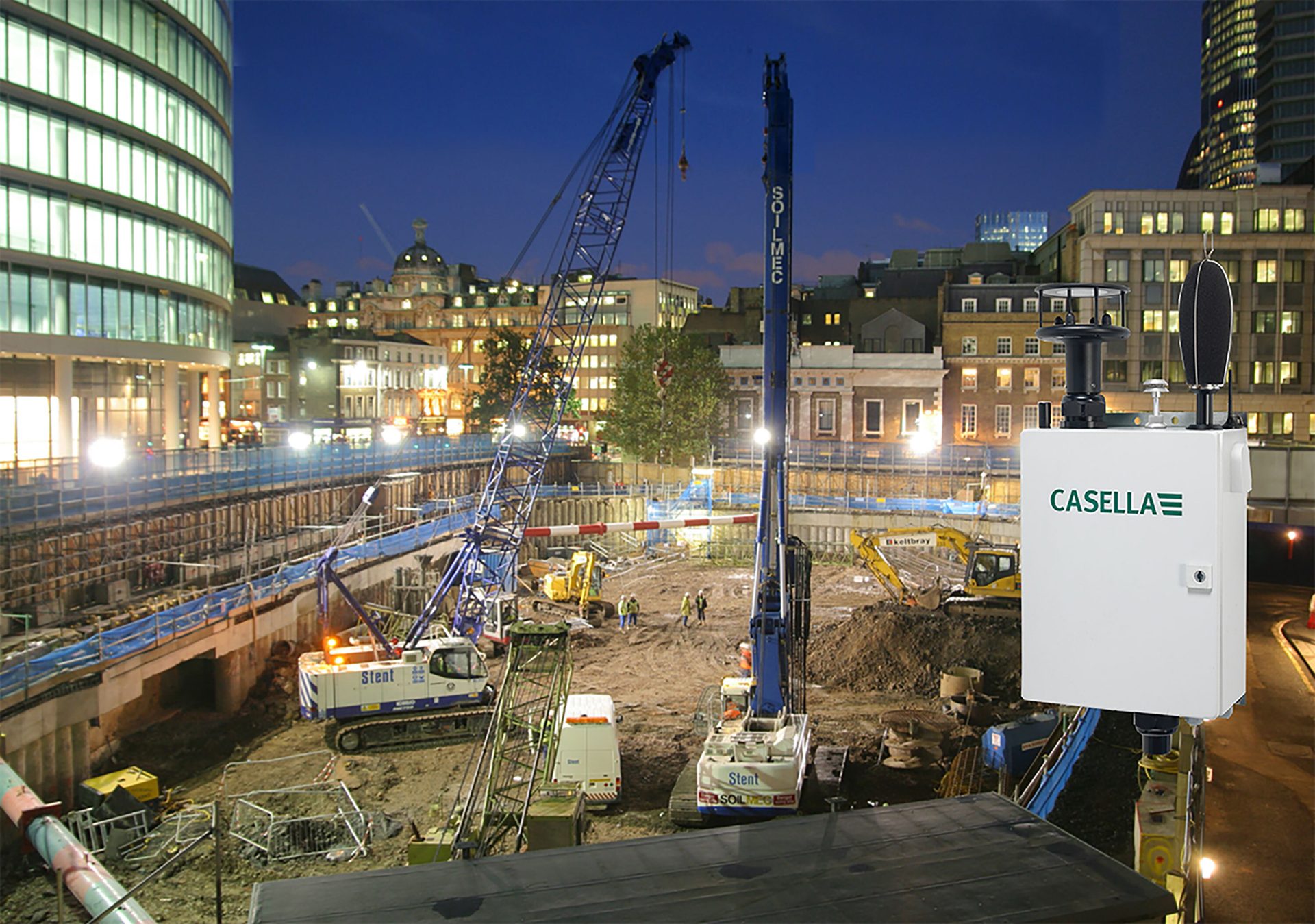 Nighttime construction site with cranes, city buildings, and a foreground Casella monitoring device.