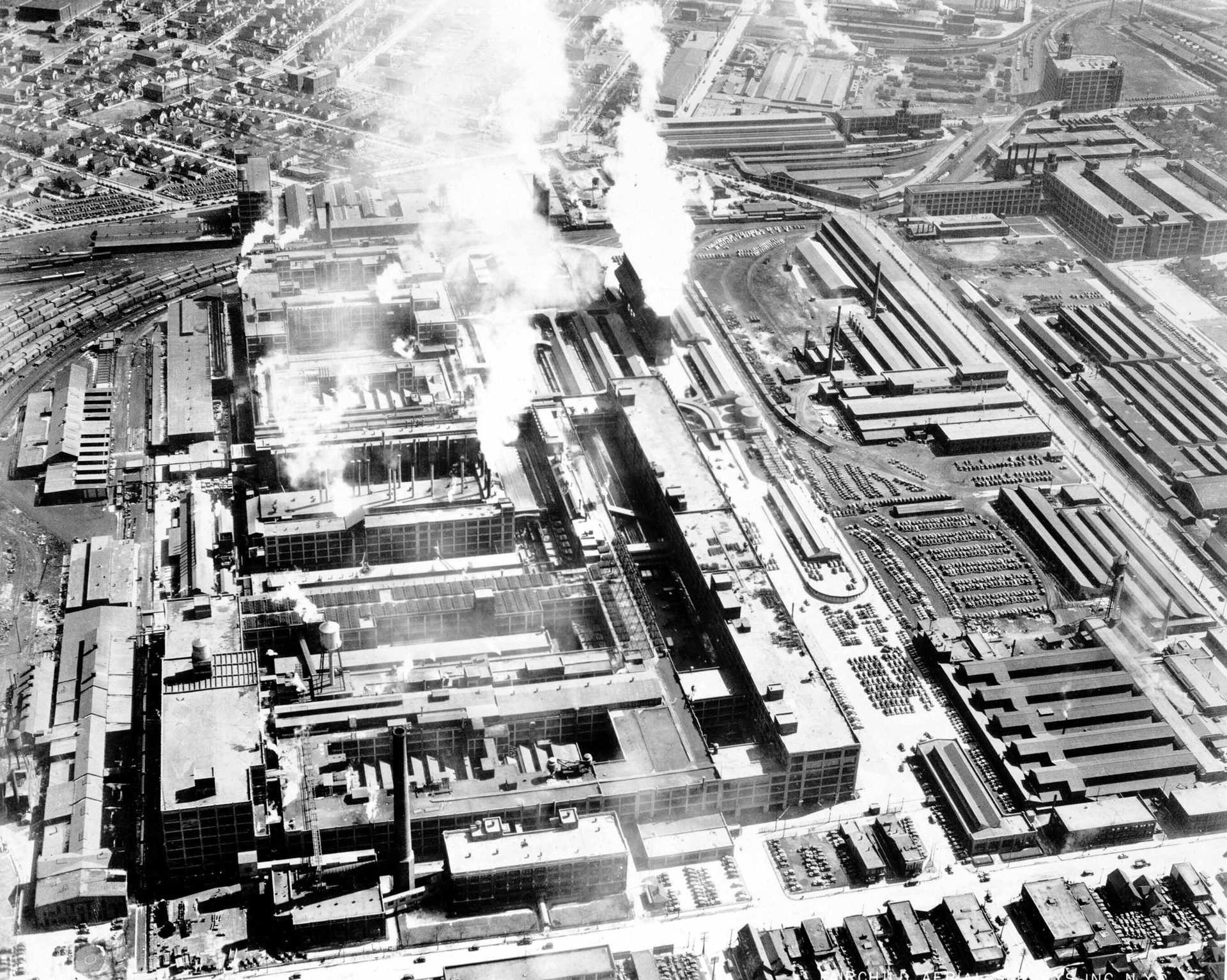 Aerial B&W of a sprawling industrial complex: smoking chimneys, train tracks, parking lots, and homes.