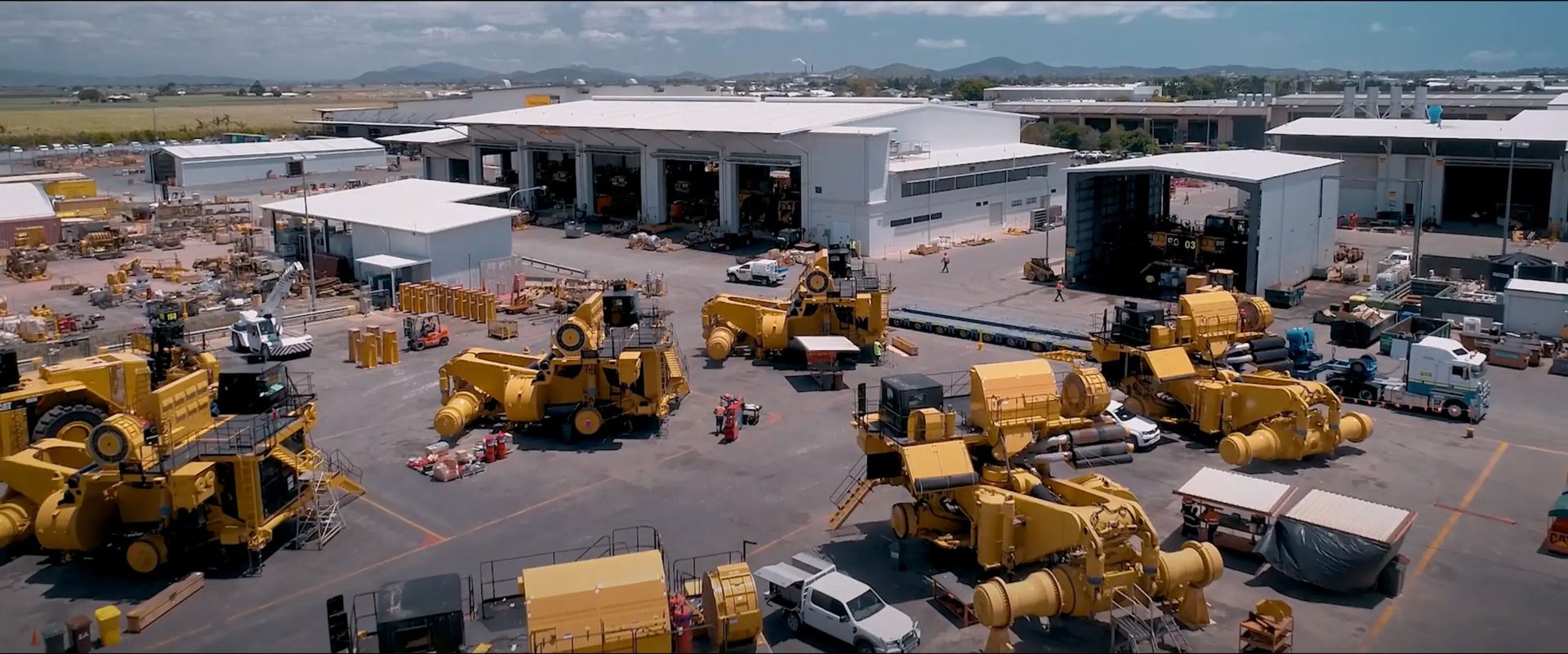 Industrial yard with huge yellow mining equipment, assembly buildings, and workers.