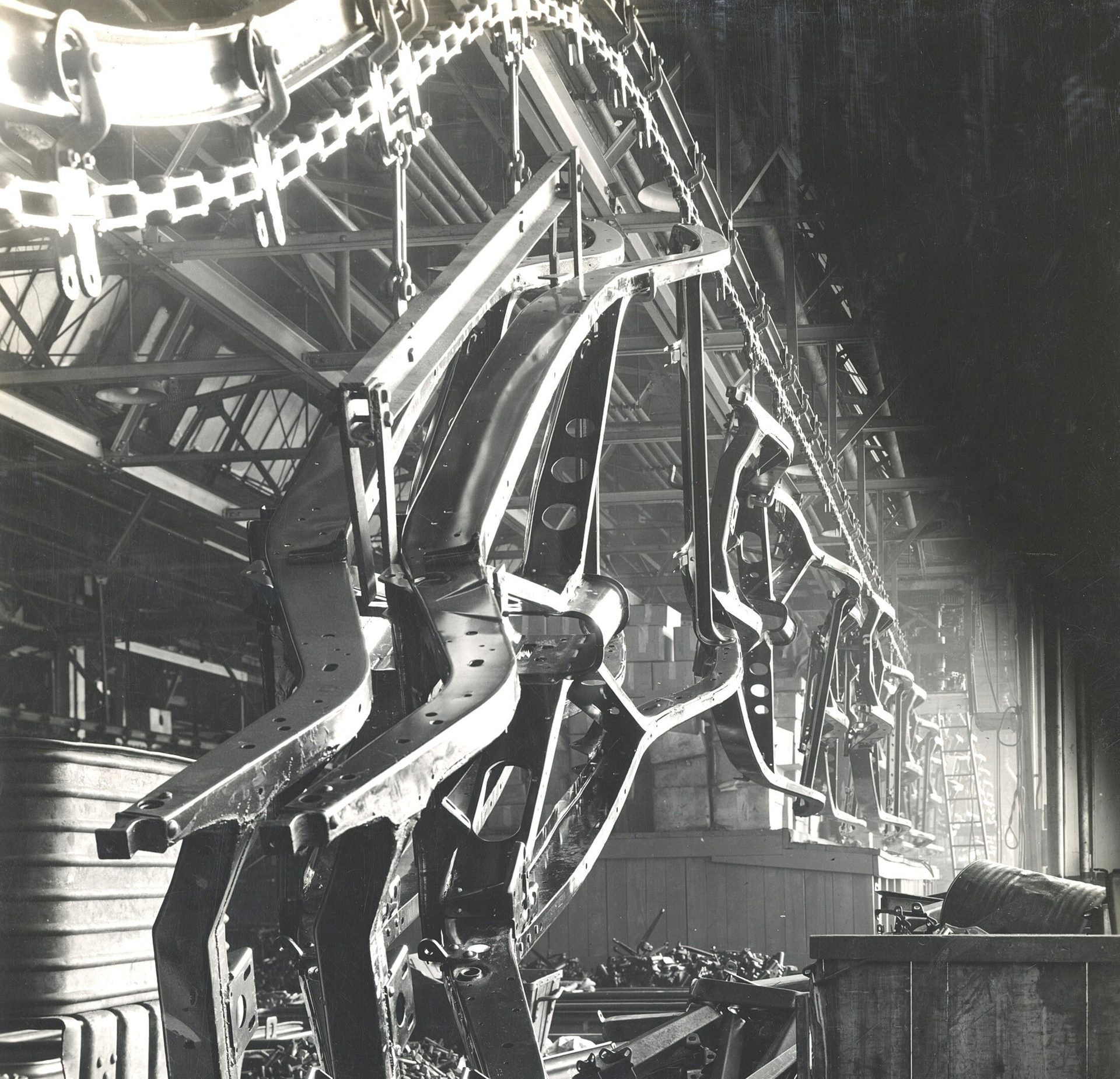 Black and white image of car chassis hanging from an overhead conveyor in a factory.