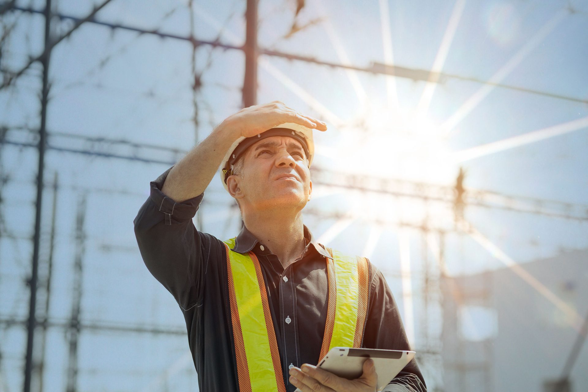 Man in hard hat, safety vest looking up, shielding eyes from sun, holding tablet at power station.