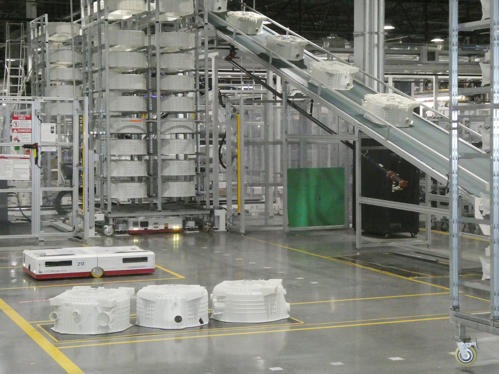 Factory floor with conveyor carrying white parts, storage racks, and an autonomous robot.