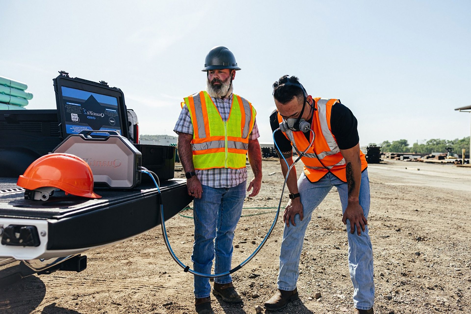 Two construction workers; one wears a respirator connected to equipment on a truck bed.