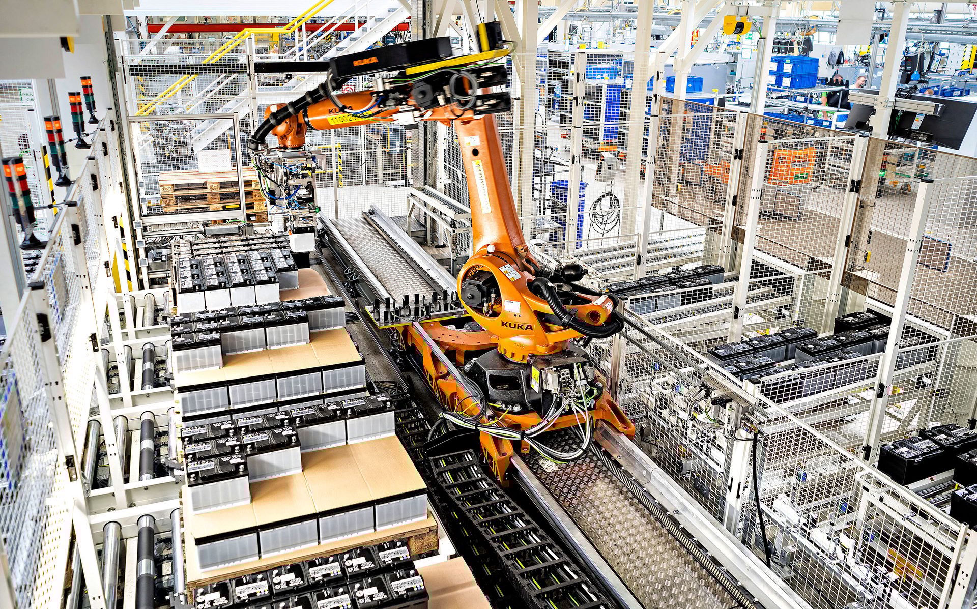Orange KUKA robot on a track in a factory, surrounded by stacks of batteries and safety cages.