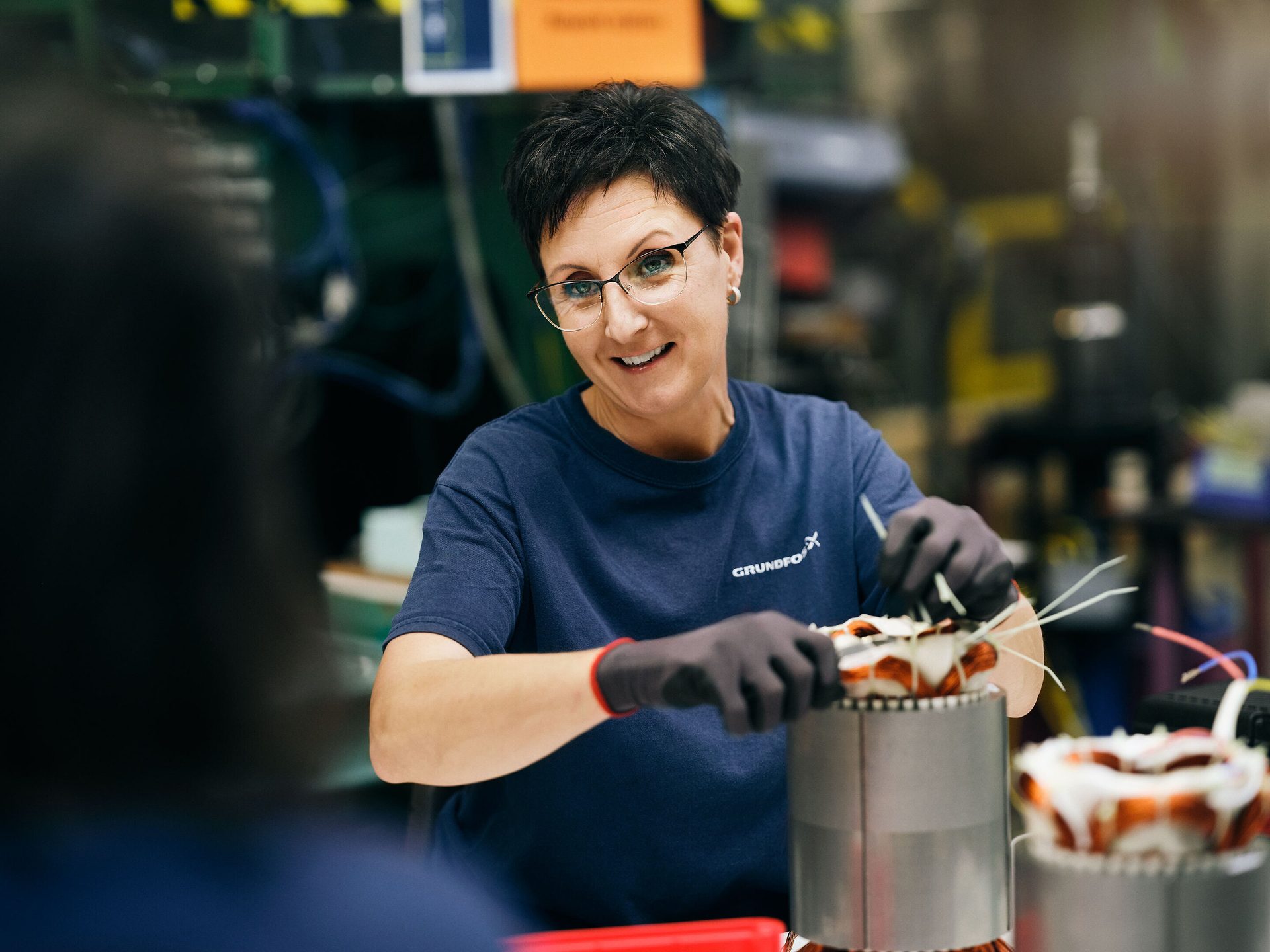Smiling woman in Grundfos shirt and gloves assembles electrical motor coils in a factory.