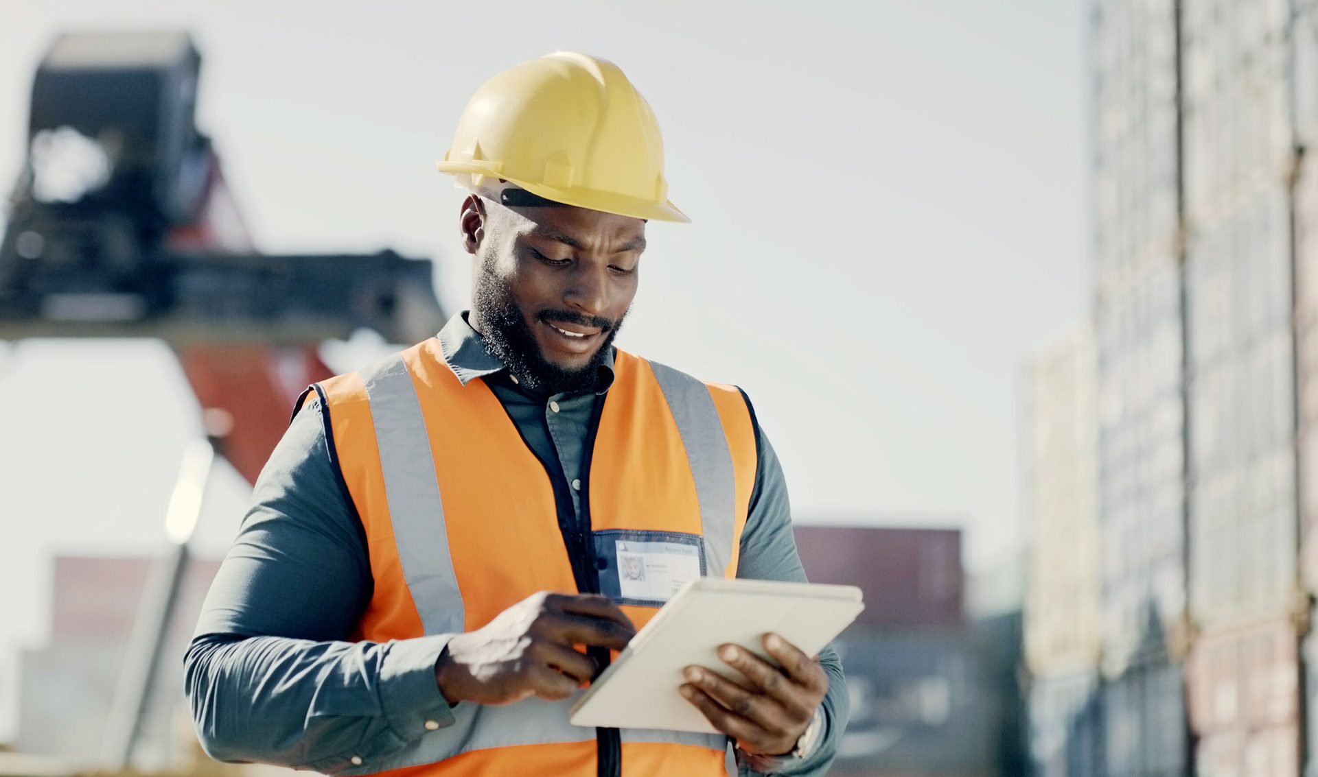 Smiling Black man in hard hat and safety vest using a tablet at a shipping yard.