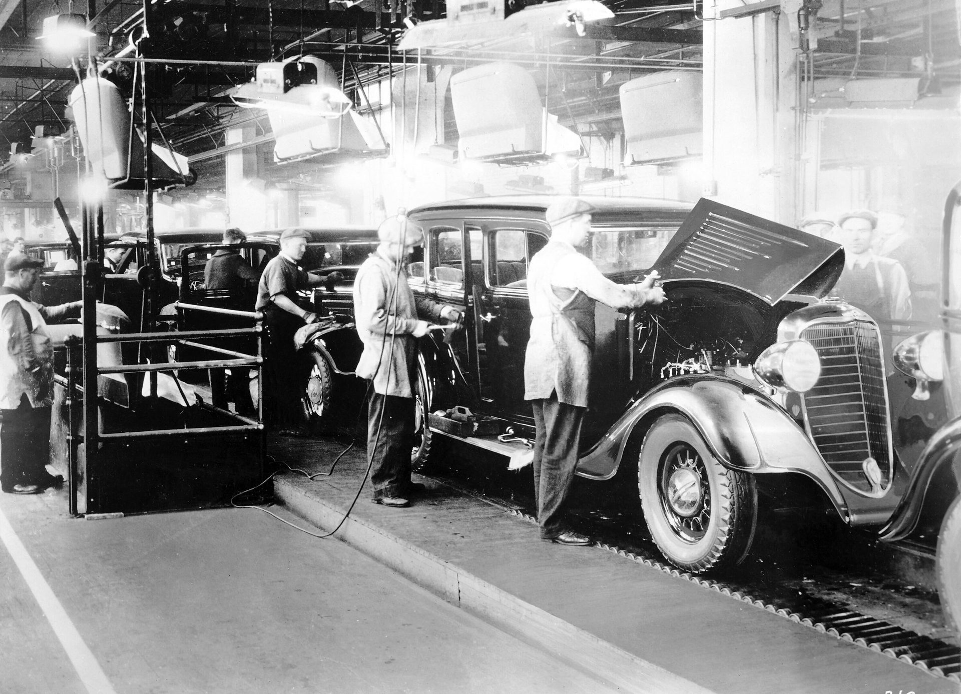 Factory workers assemble vintage cars on a production line under bright lights.