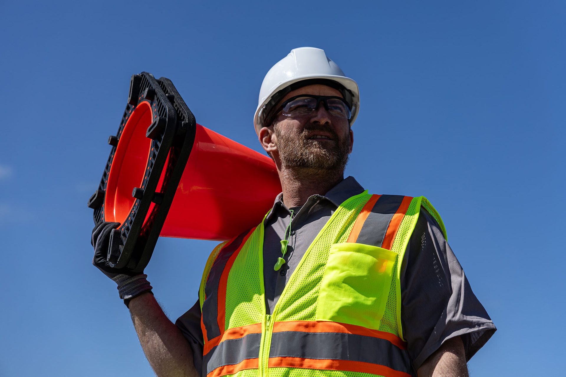 Worker in hard hat, safety vest, carries orange traffic cone on shoulder.