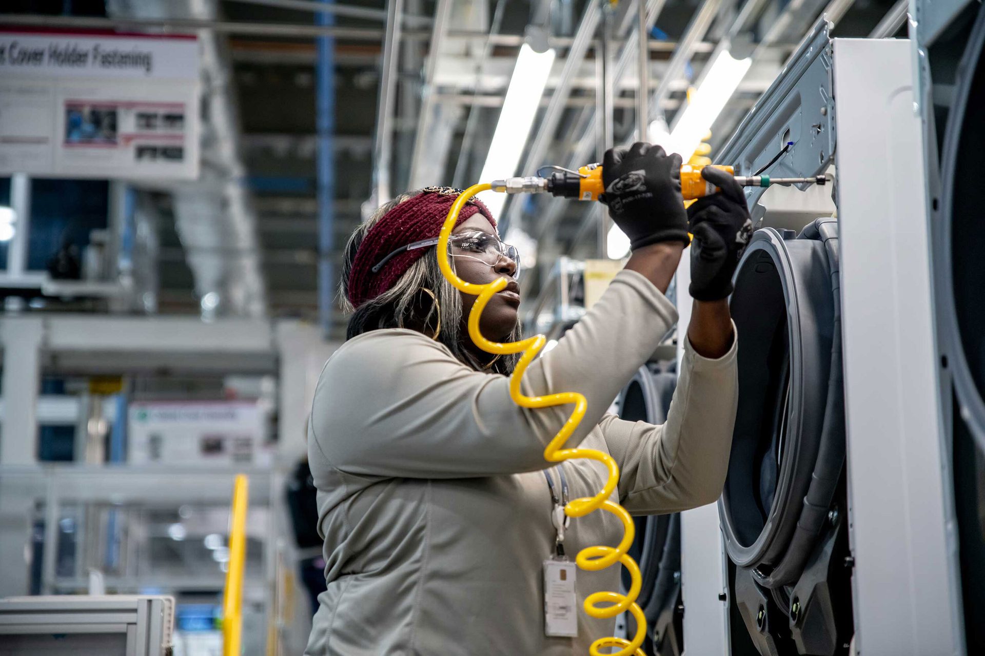 Black woman in a factory assembling an appliance with a power tool, wearing safety glasses.