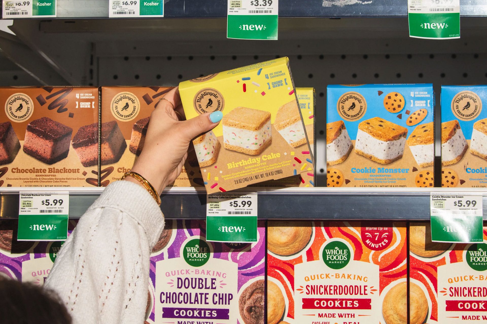 A hand selects a "Birthday Cake" ice cream sandwich box from a supermarket freezer shelf.