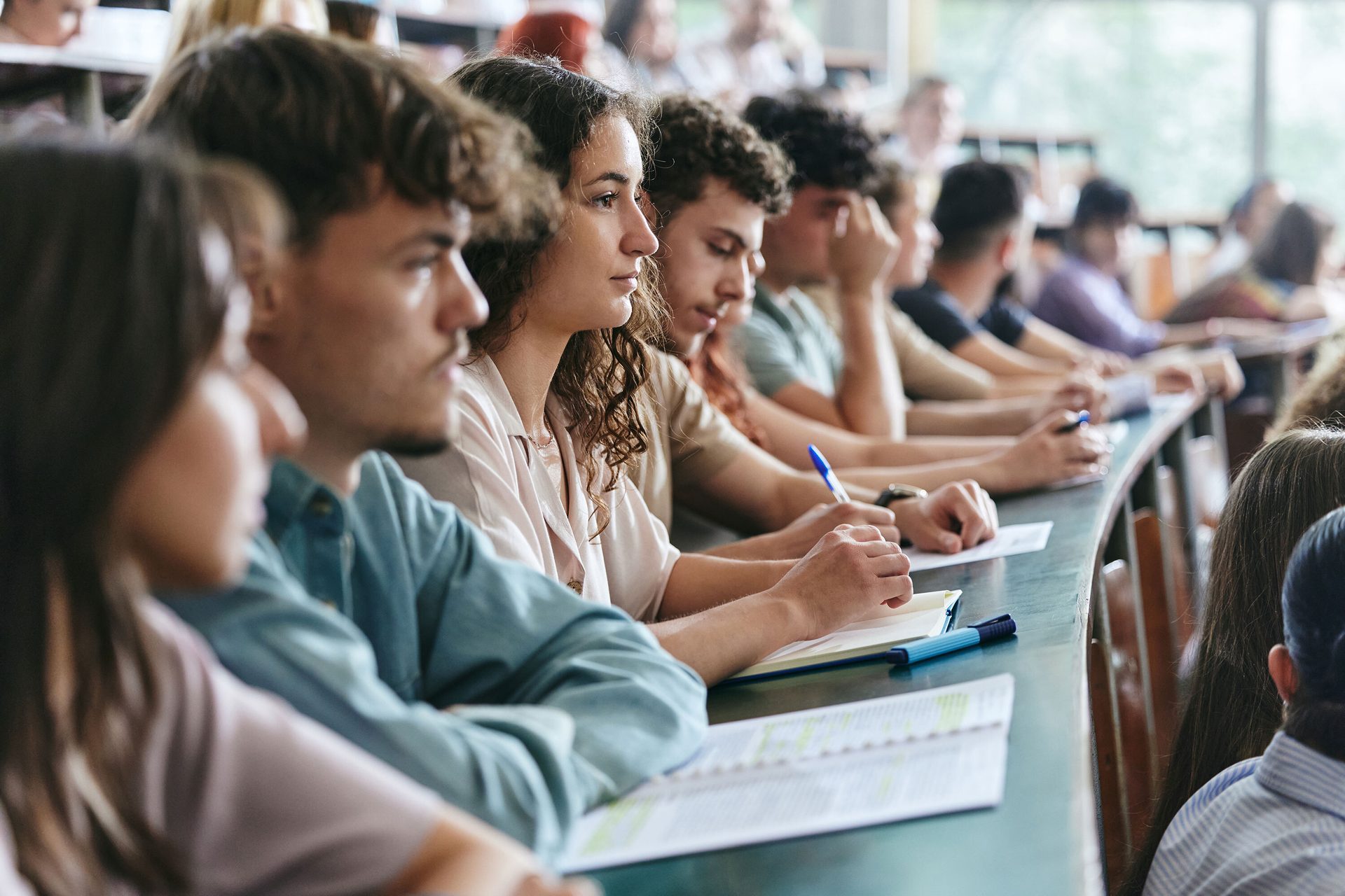 Diverse students in a lecture hall are focused, some taking notes.
