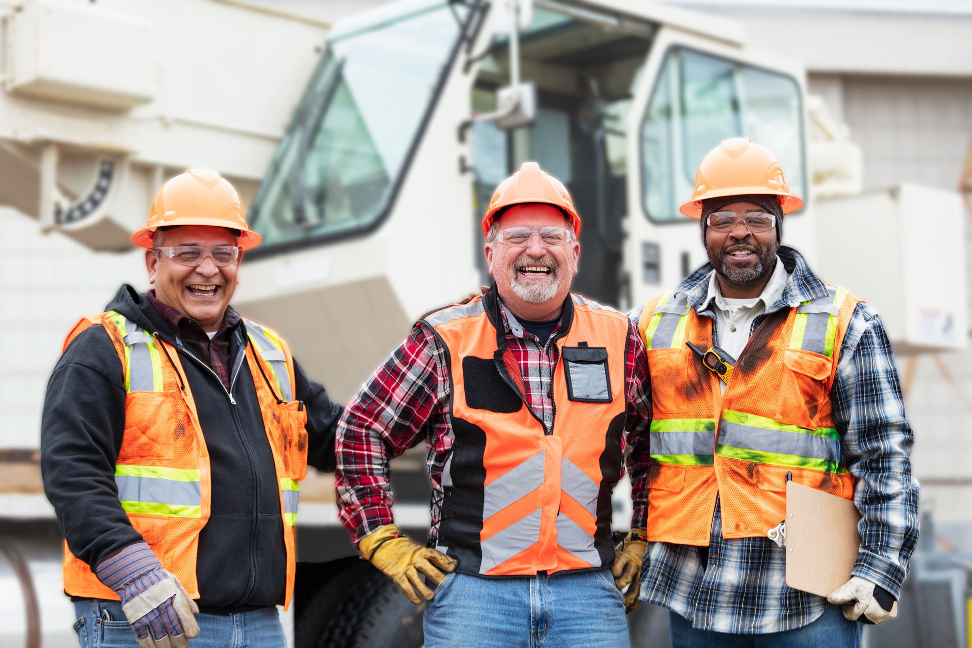 Three smiling construction workers in hard hats and safety vests stand in front of heavy machinery.