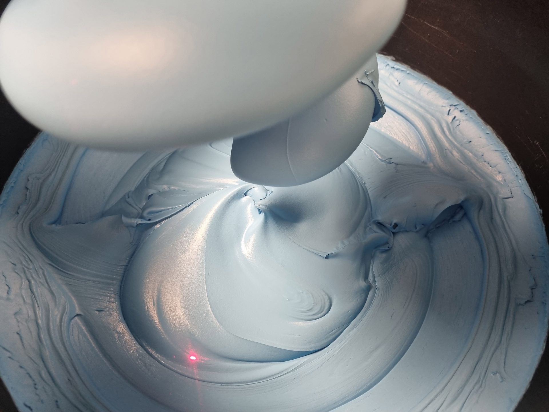 Light blue glossy cream being mixed in a bowl, with a mixer paddle and a red laser dot.