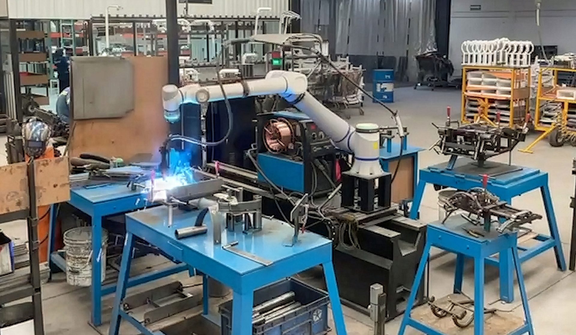 A white robotic arm performs welding on a metal piece with bright sparks in an industrial workshop.