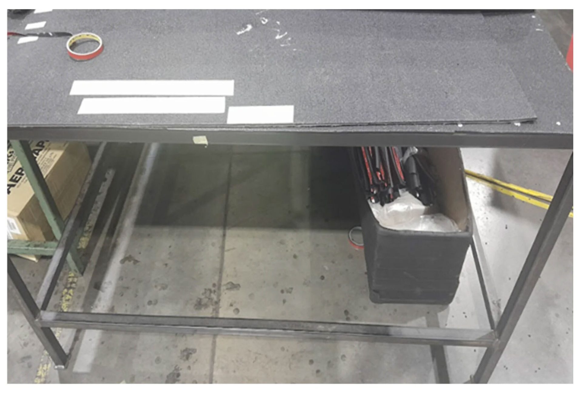 A grey workbench with white strips, tape, a metal frame below, and a black container on a concrete floor.