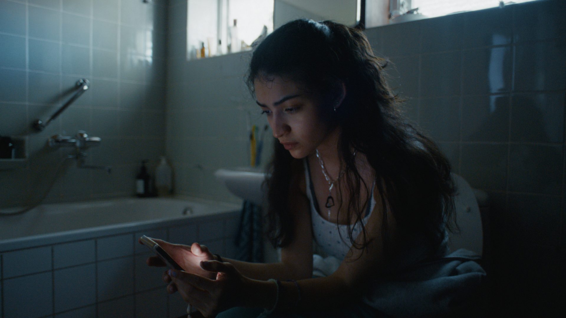 Young woman in a dimly lit bathroom, engrossed in her glowing smartphone.