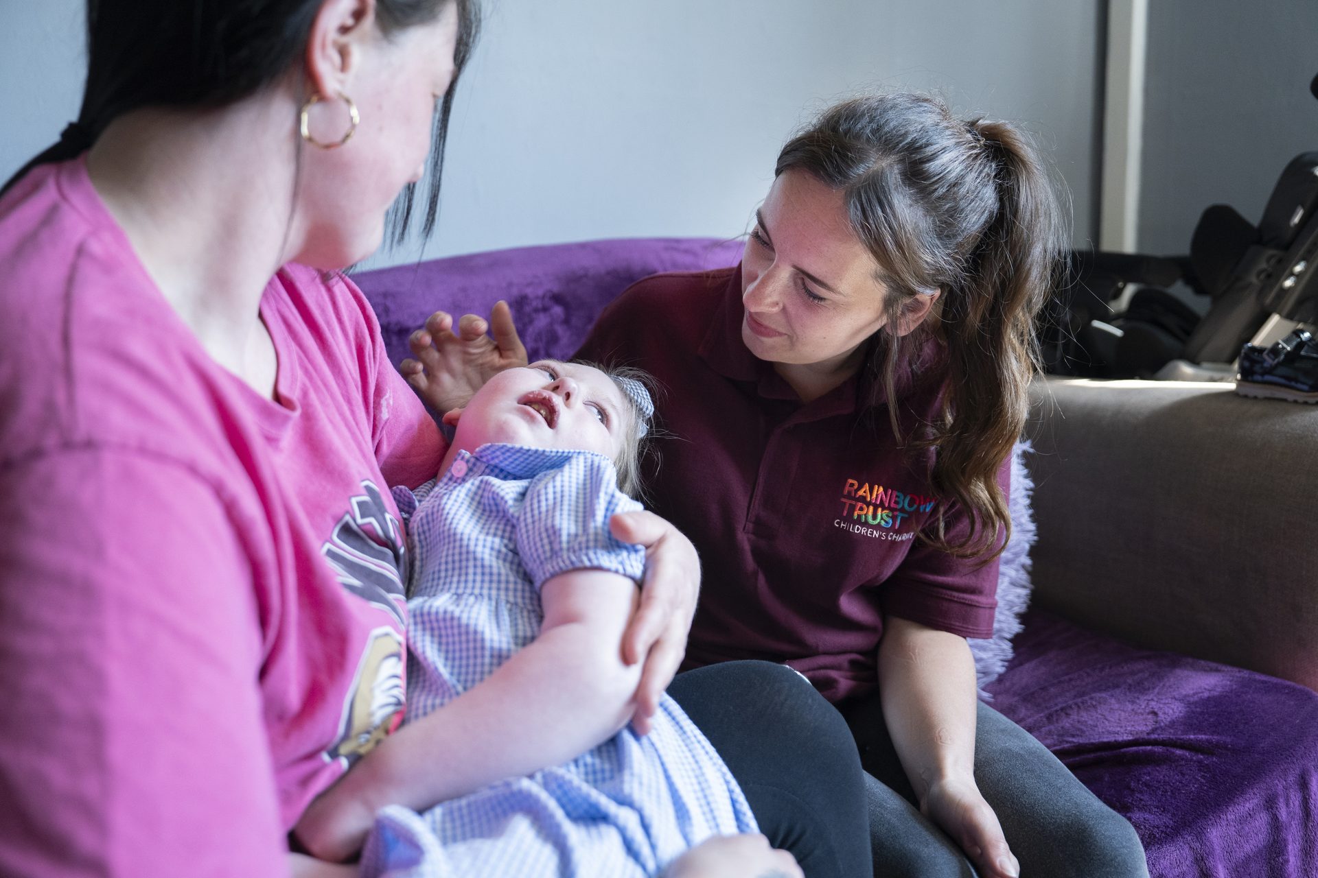 A caregiver holds a child, while a Rainbow Trust staff member smiles at them on a couch.