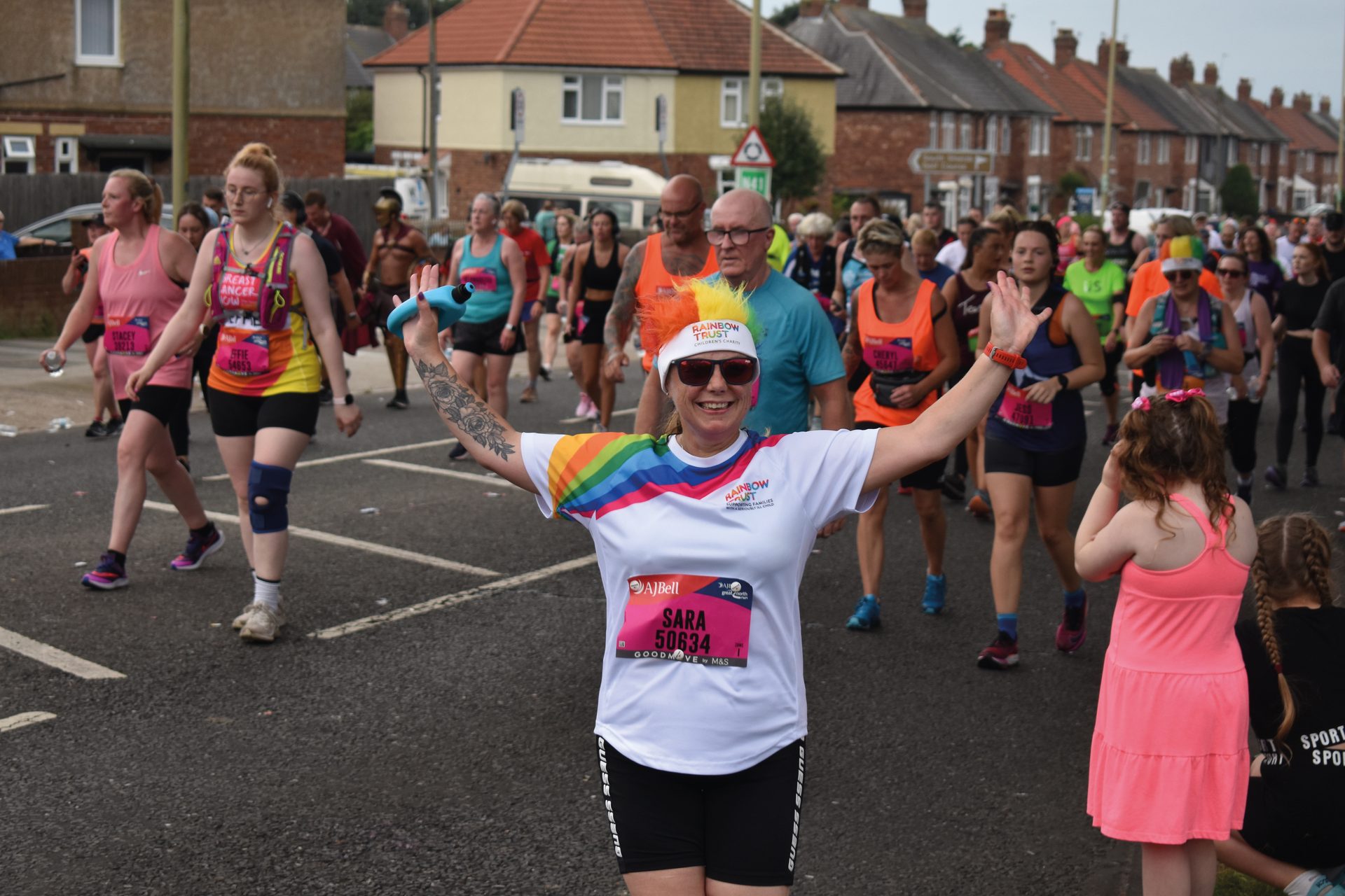 Woman in rainbow outfit with arms spread, smiling at a race with a crowd behind her.
