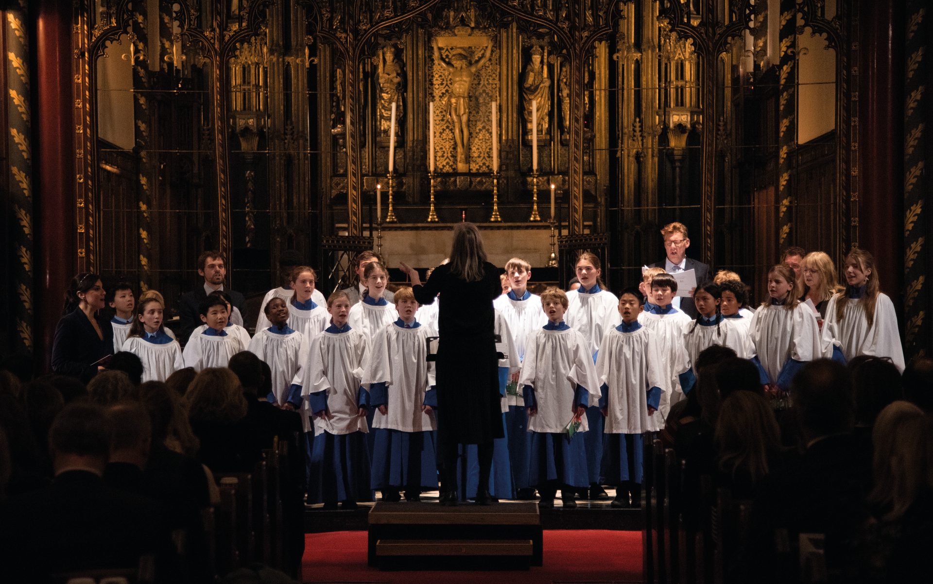 A conductor leads a children's choir in white and blue robes performing in a grand, ornate church.