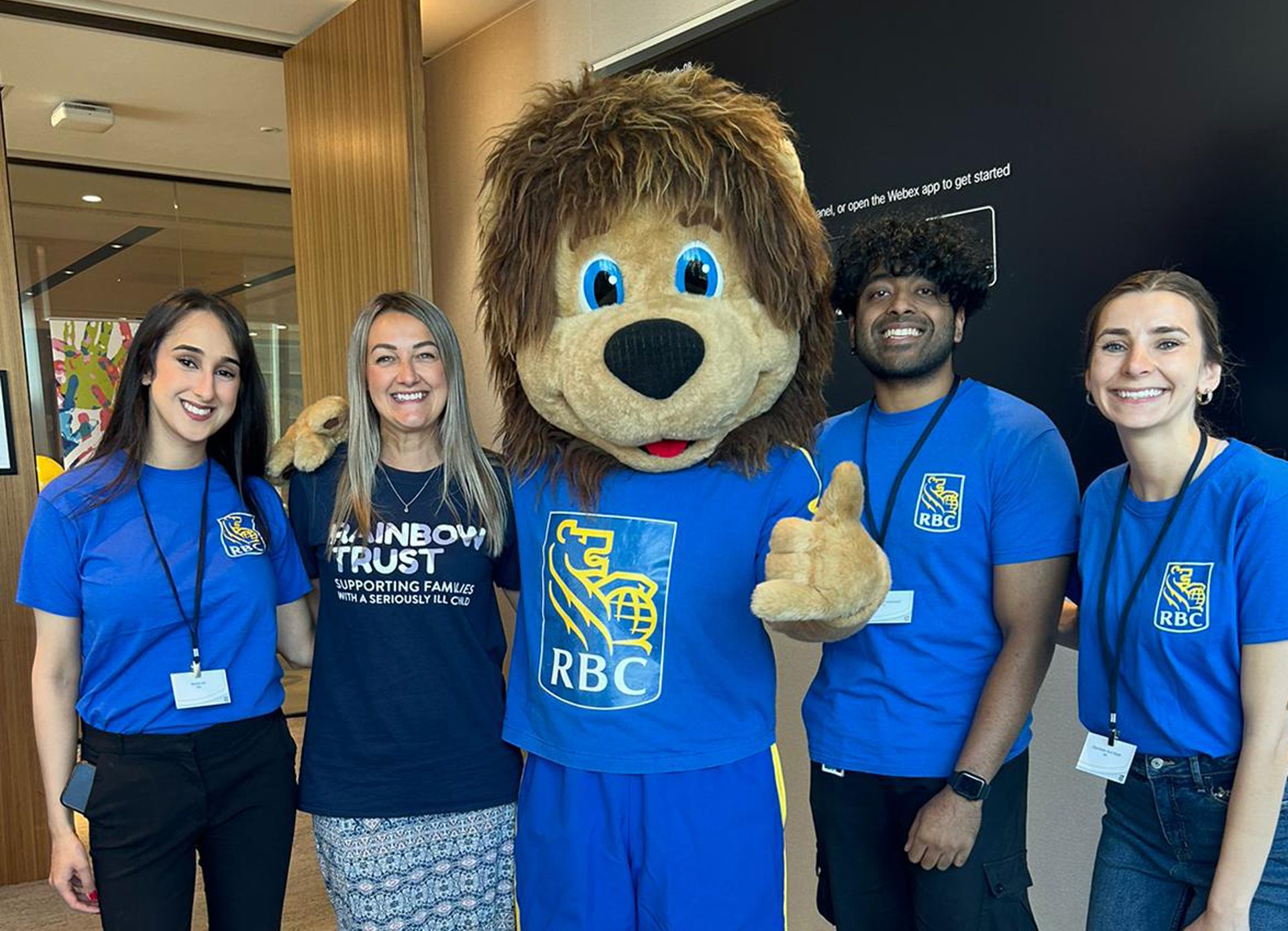 Smiling group: 4 people & lion mascot. Most in blue RBC shirts, one in Rainbow Trust shirt.