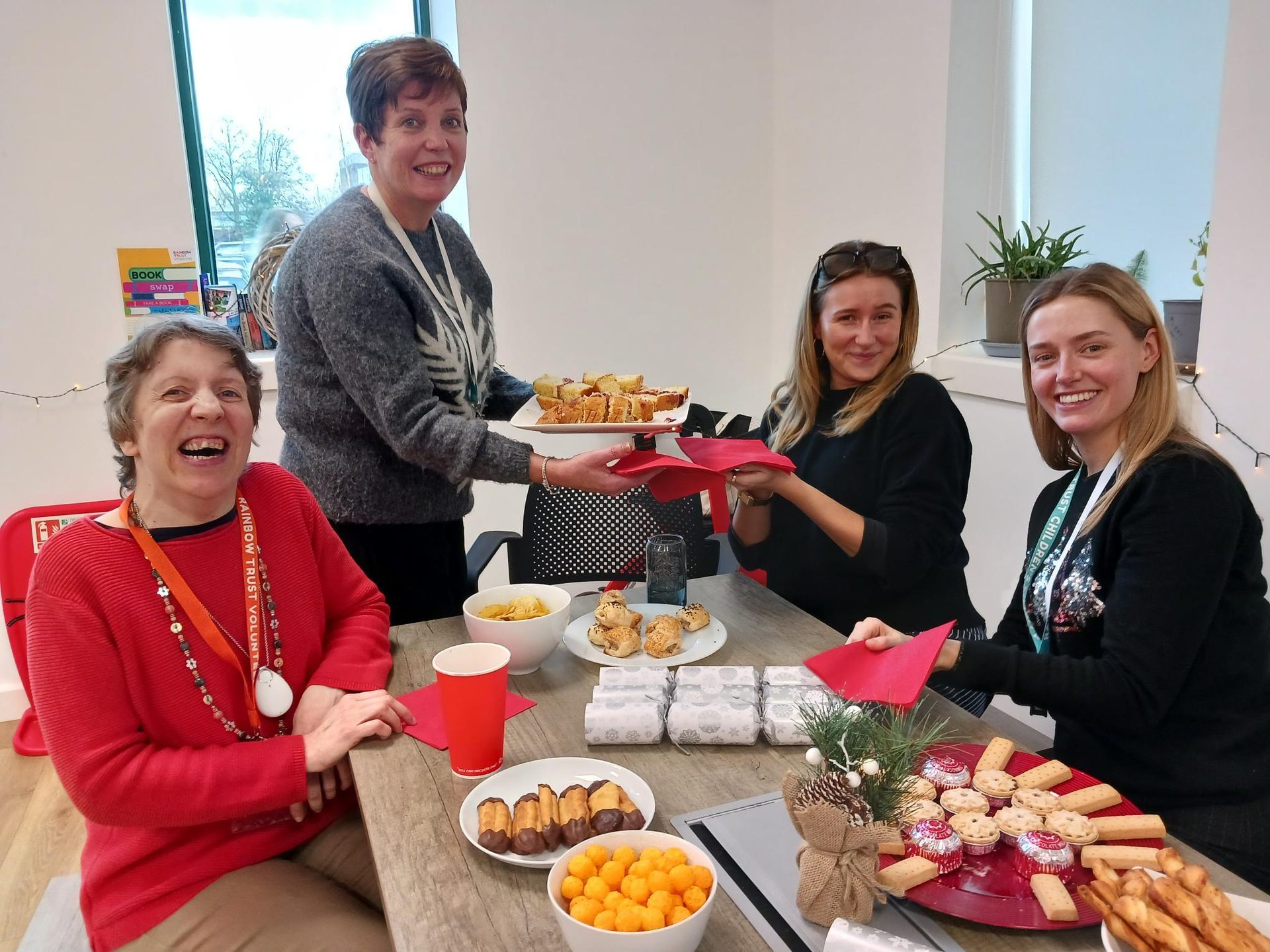 Four women happily gathered around a table with snacks; one serves food.