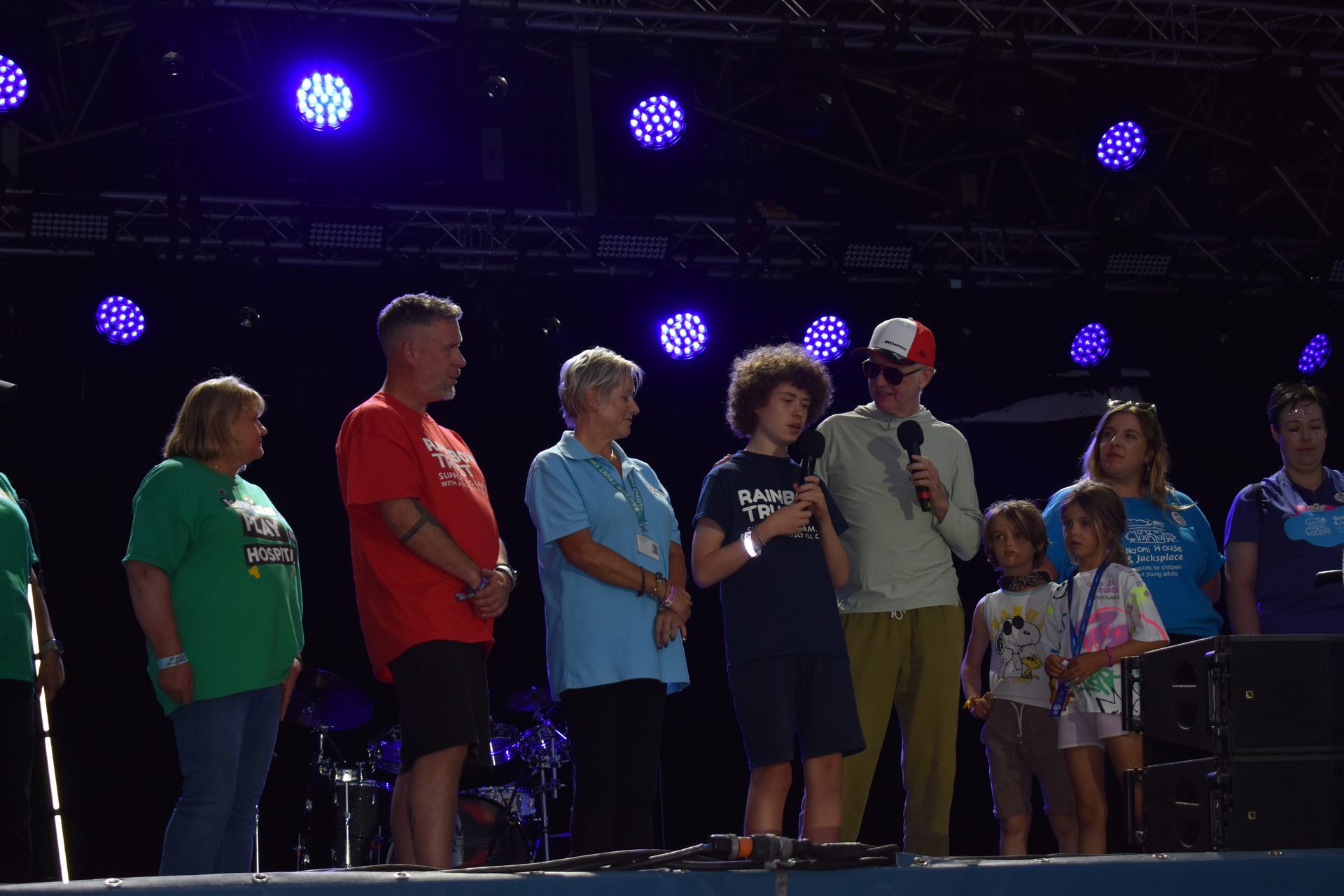 Group of adults and children on a stage under blue lights, some speaking into microphones.