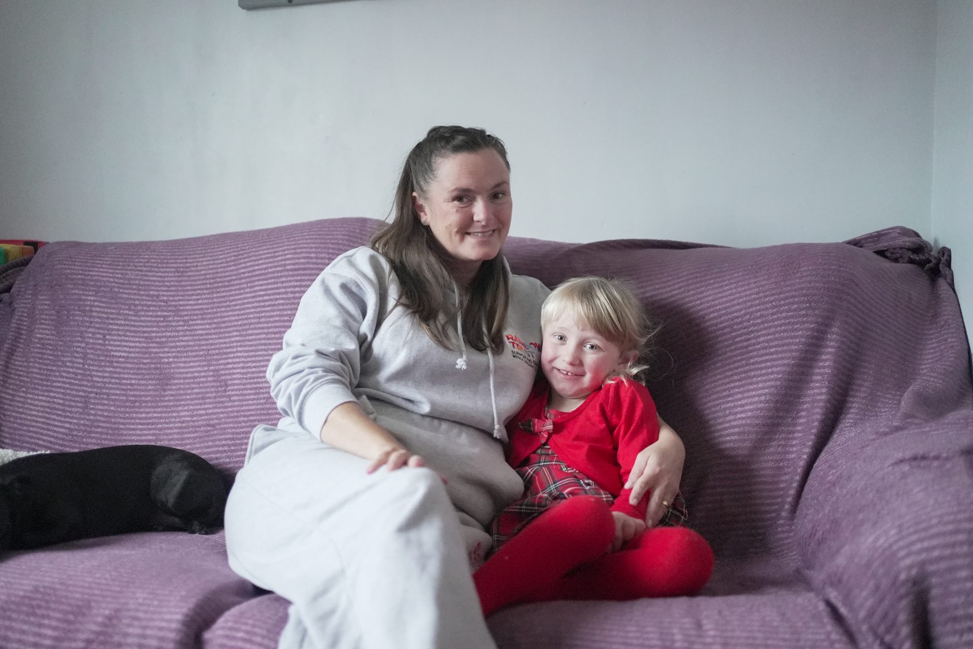 A woman and child smile on a purple couch, with a dark dog partially visible on the left.