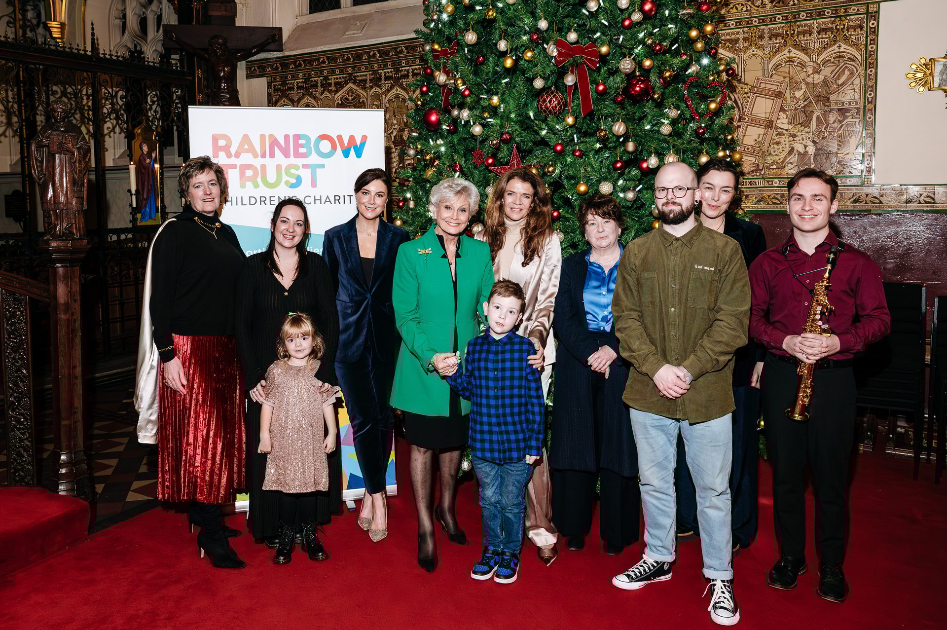A group of adults and two children pose in front of a Christmas tree and "Rainbow Trust Children's Charity" banner.