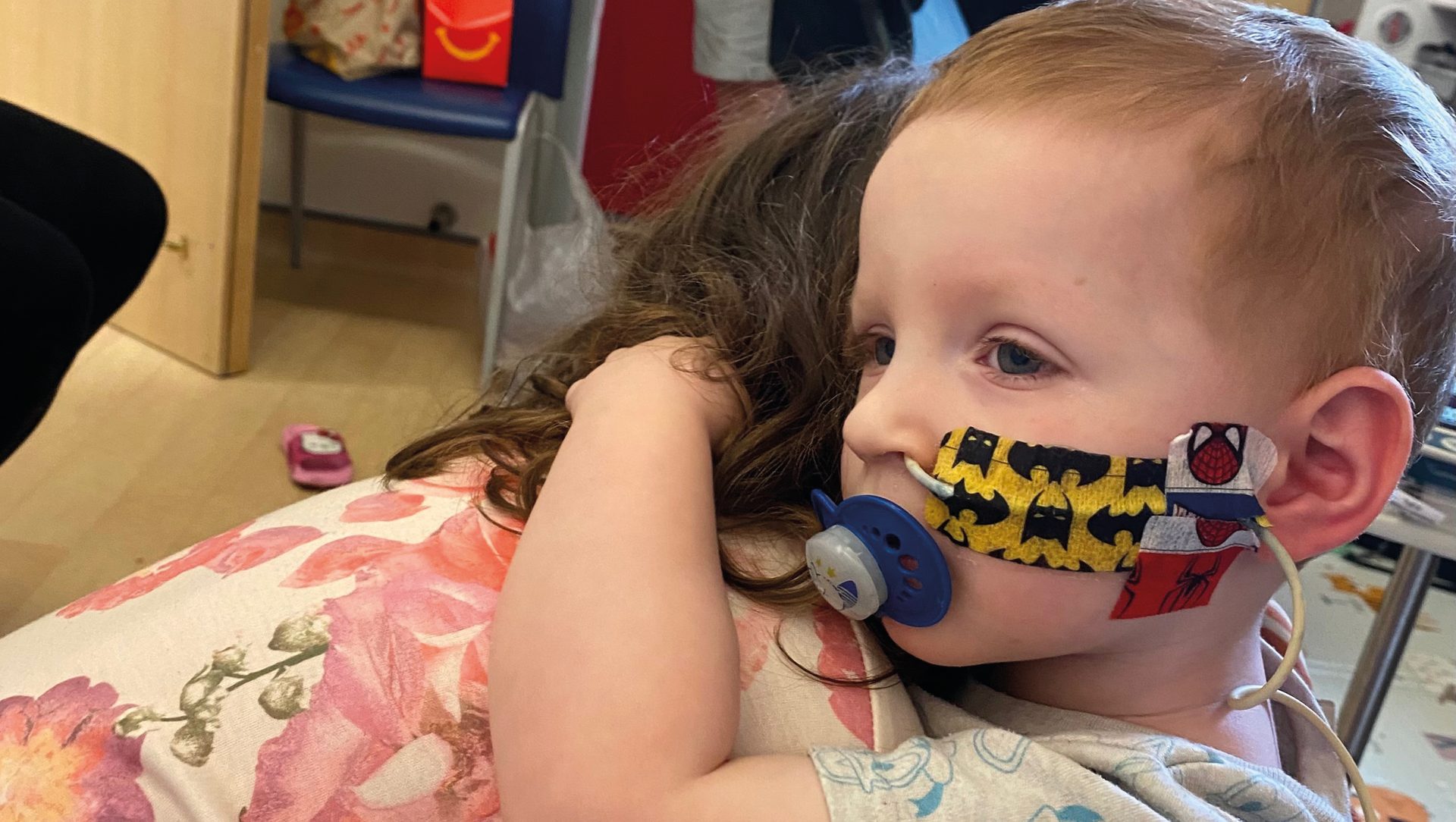 Young child with decorated medical tubes and pacifier, embraced by an adult.