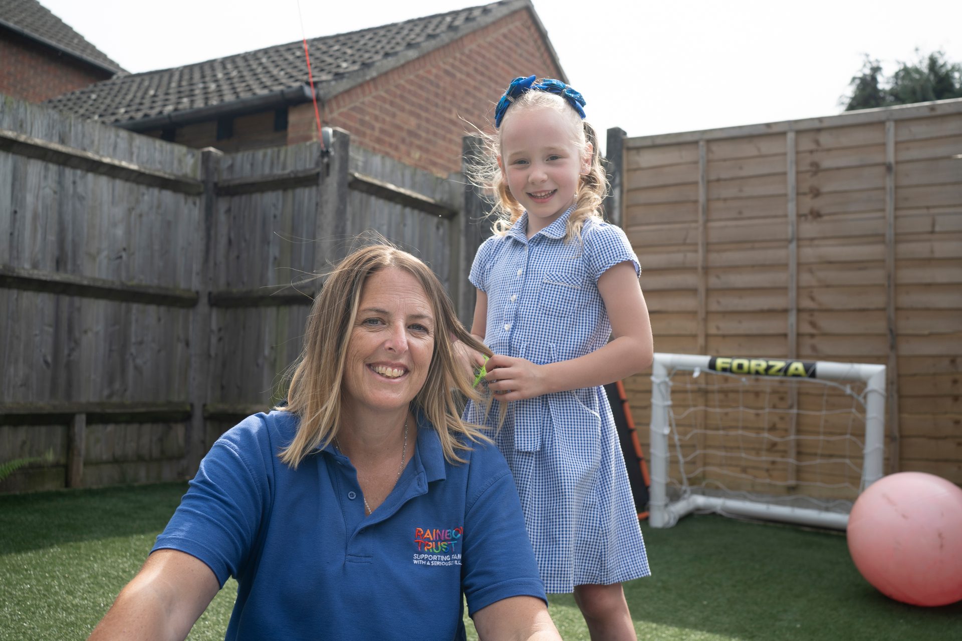 Smiling woman in 'Rainbow Trust' shirt, with a girl playfully styling her hair in a backyard.