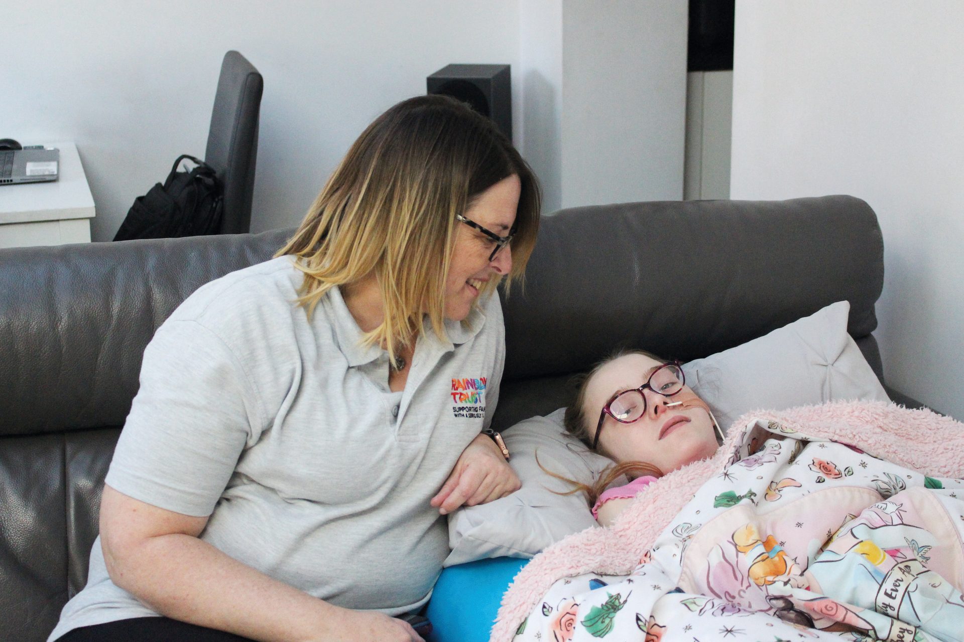 Woman in Rainbow Trust shirt smiles at girl with nasal cannula on couch.