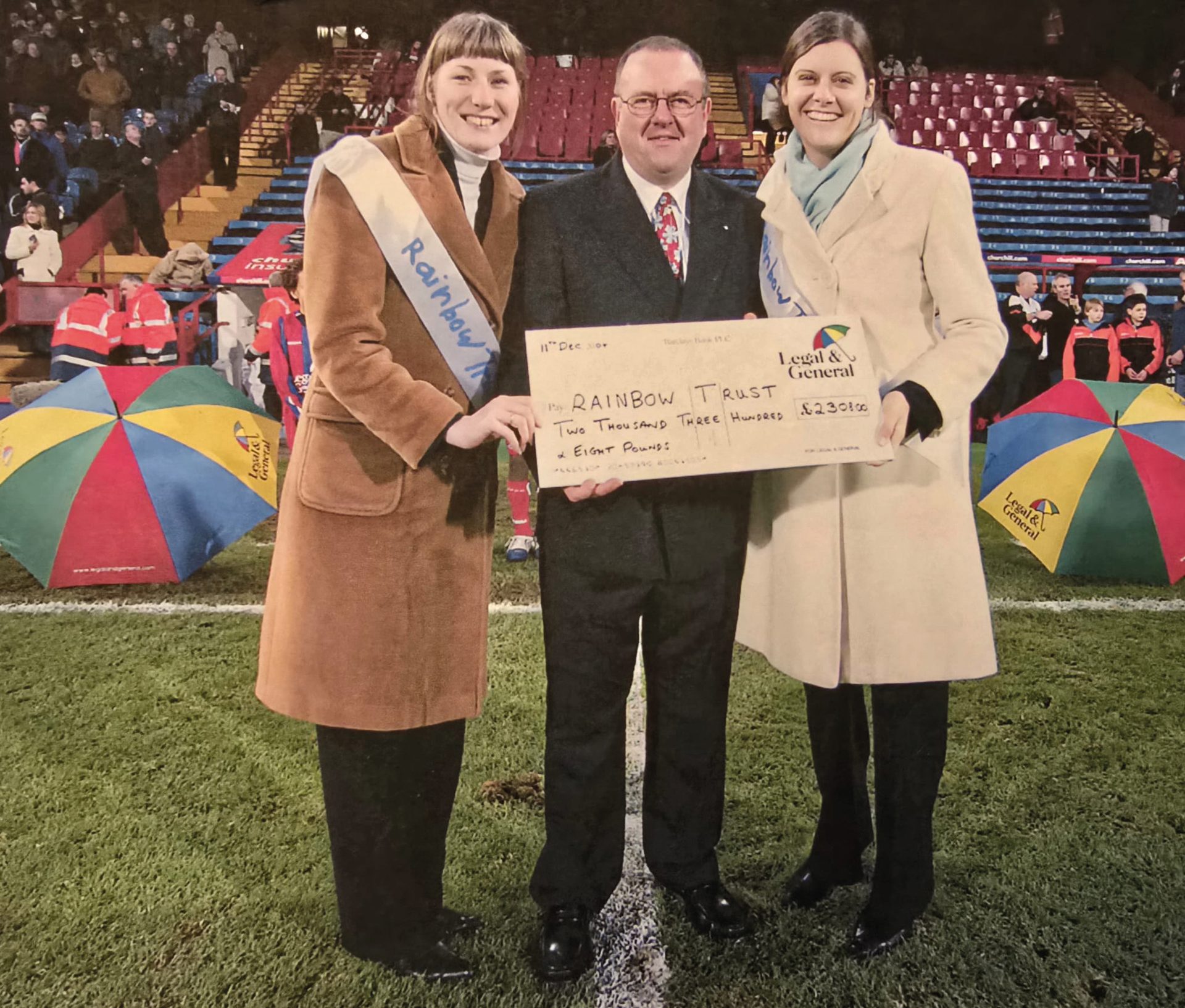 Three people, two women and a man, hold a large check for Rainbow Trust on a football field.