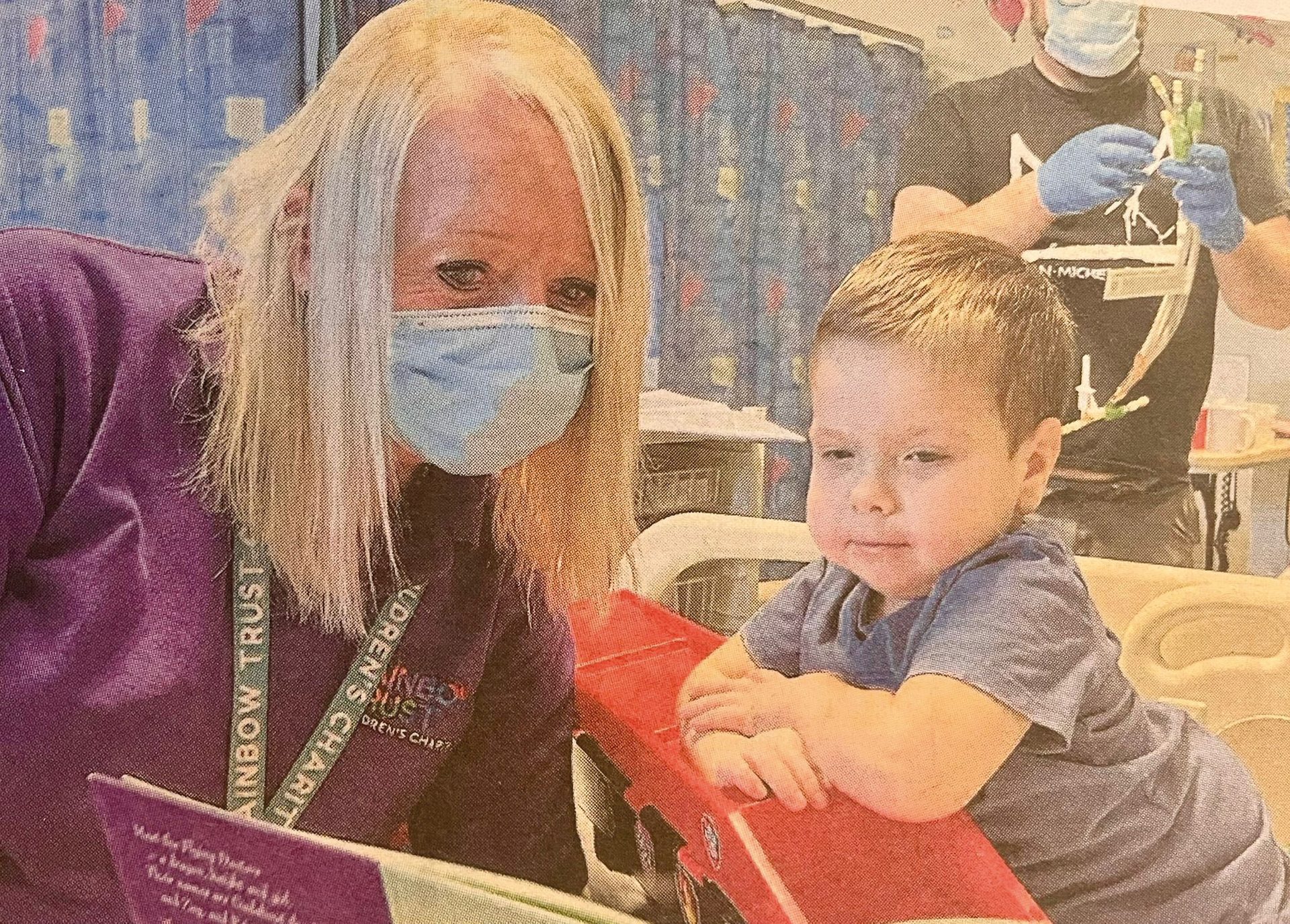 A masked woman reads a book to a young boy leaning on a red toy in a hospital room.