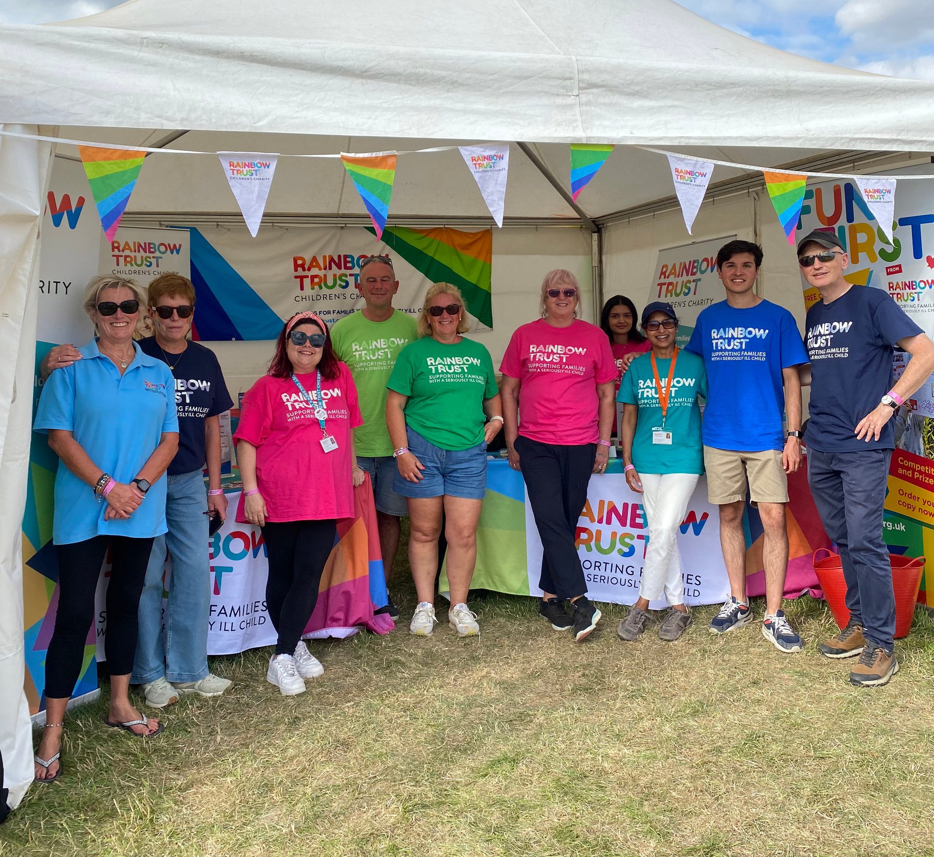 Rainbow Trust Children's Charity volunteers pose in front of their branded tent at an outdoor event.