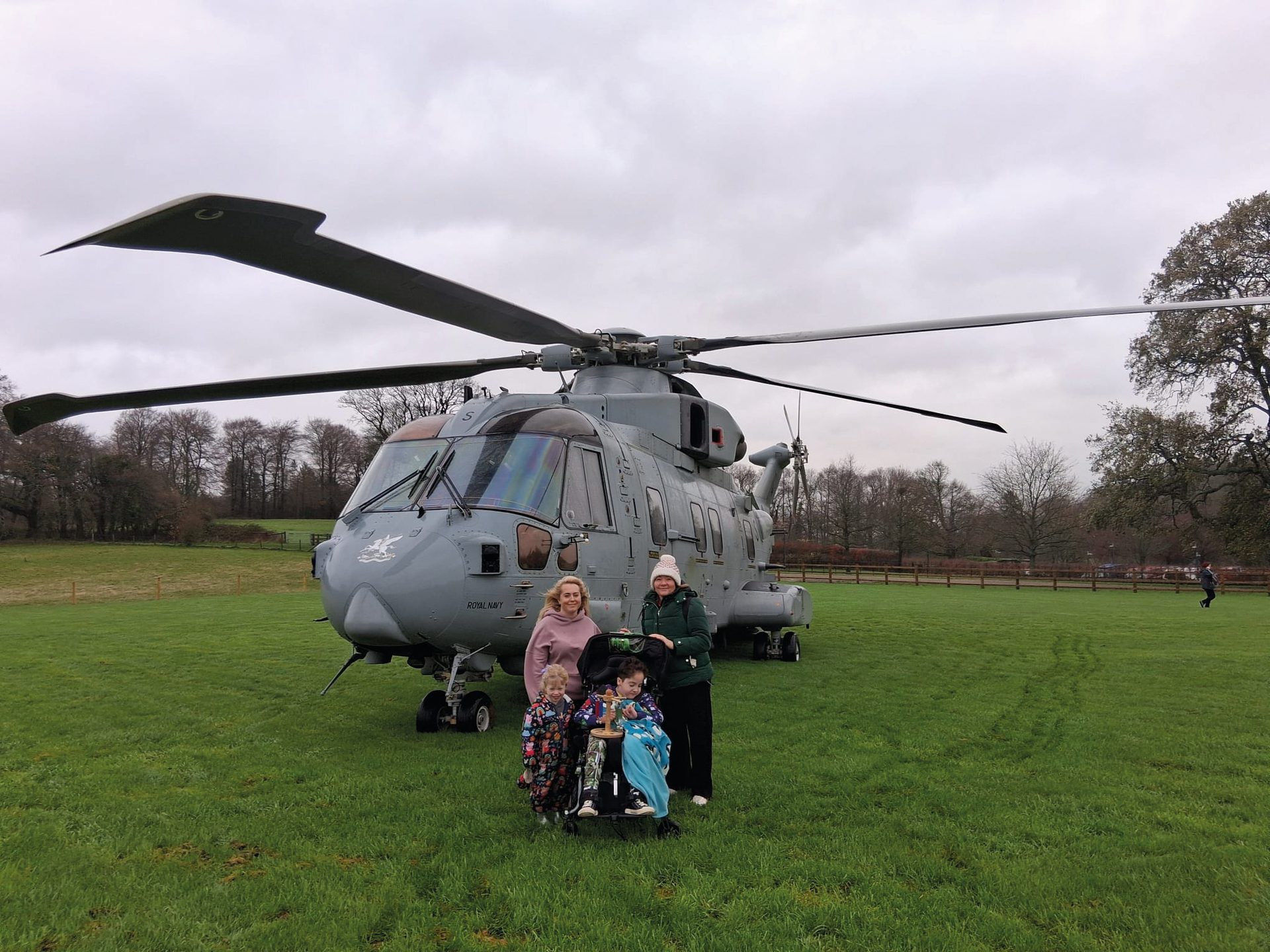 A family poses in front of a large grey Royal Navy helicopter on a grassy field under a cloudy sky.