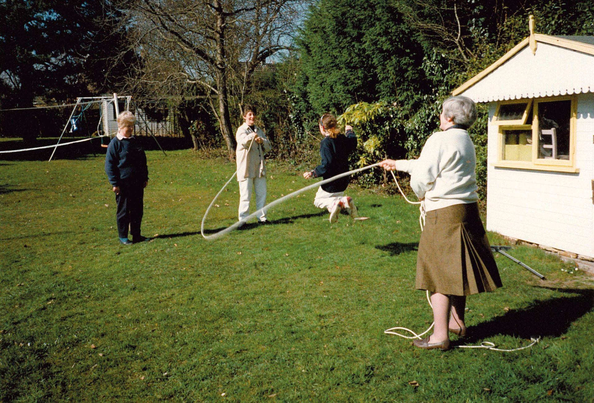 Three women and a girl jump roping in a sunny backyard with a shed and trees.