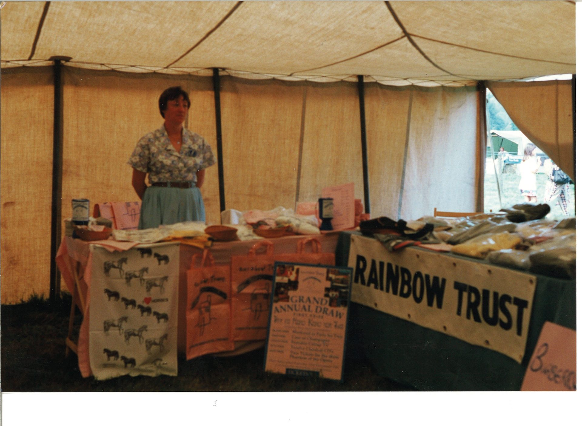 A woman stands behind a table with goods inside a tent, with a 'Rainbow Trust' sign visible.