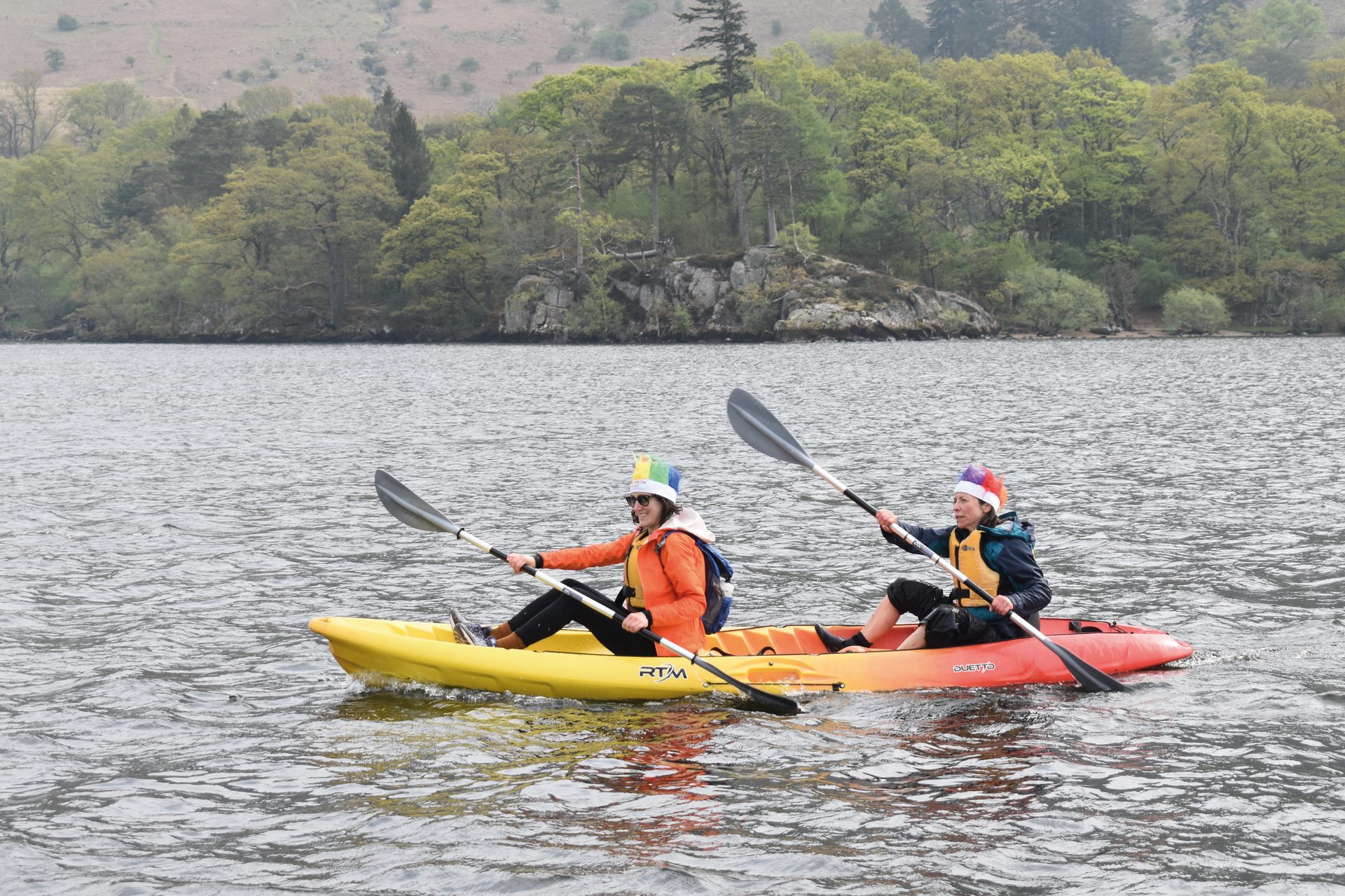 Two people in colorful hats paddle kayaks on a lake with a treelined shore in the background.