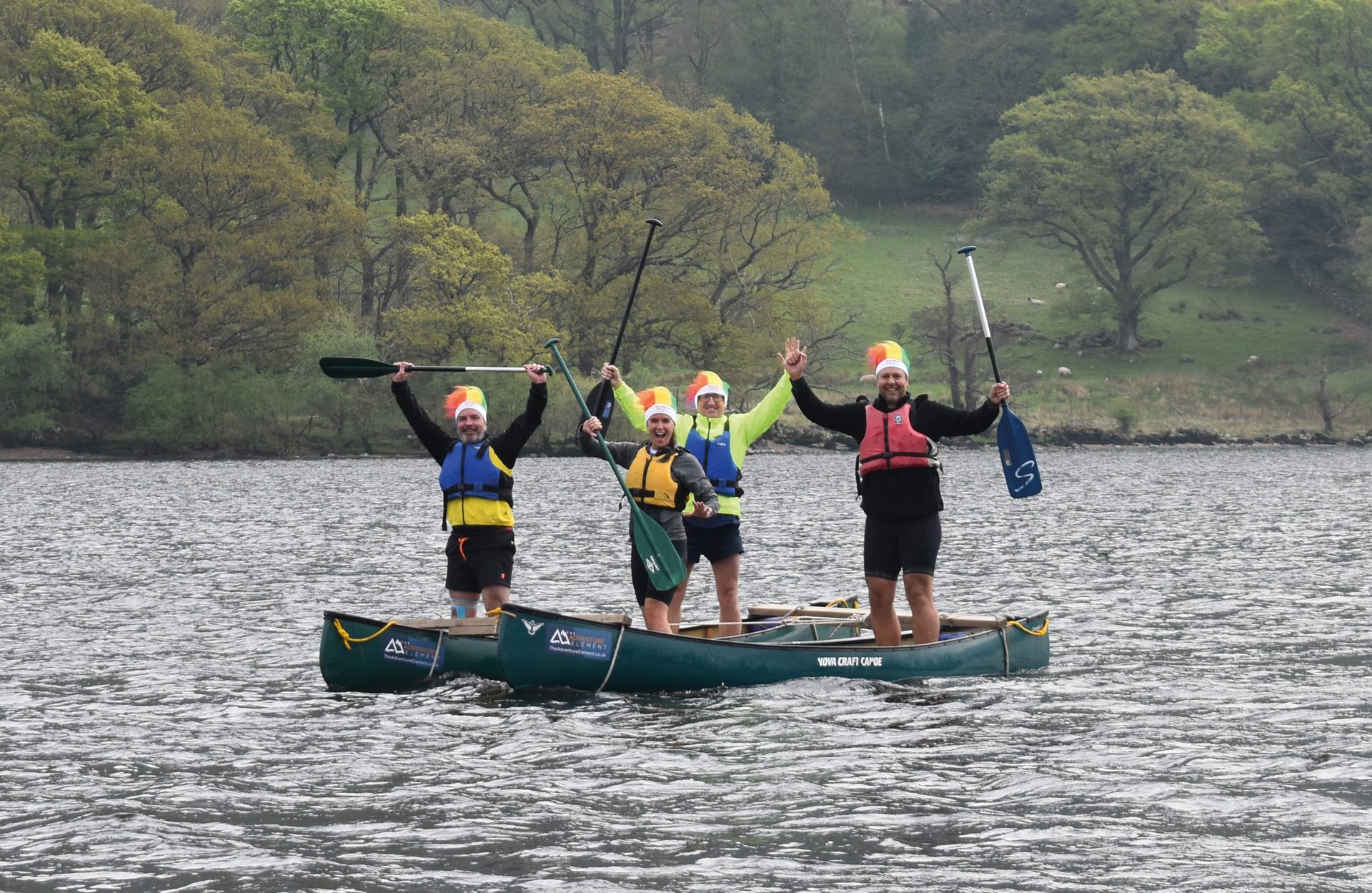 Four happy people in rainbow hats stand in canoes on a lake, arms raised in celebration.