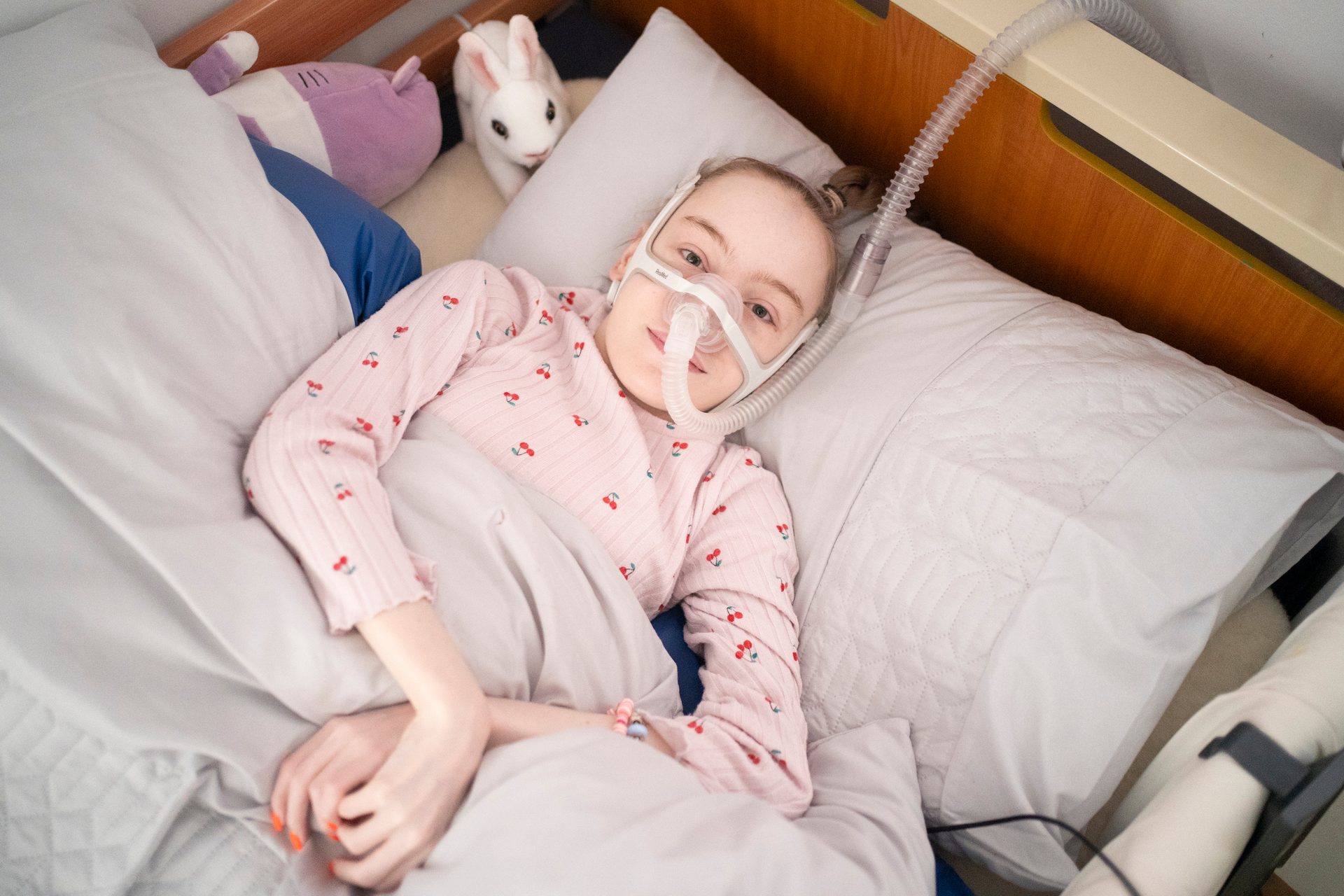 Young person in bed with a medical mask, pink cherry shirt, and a white rabbit plushie.