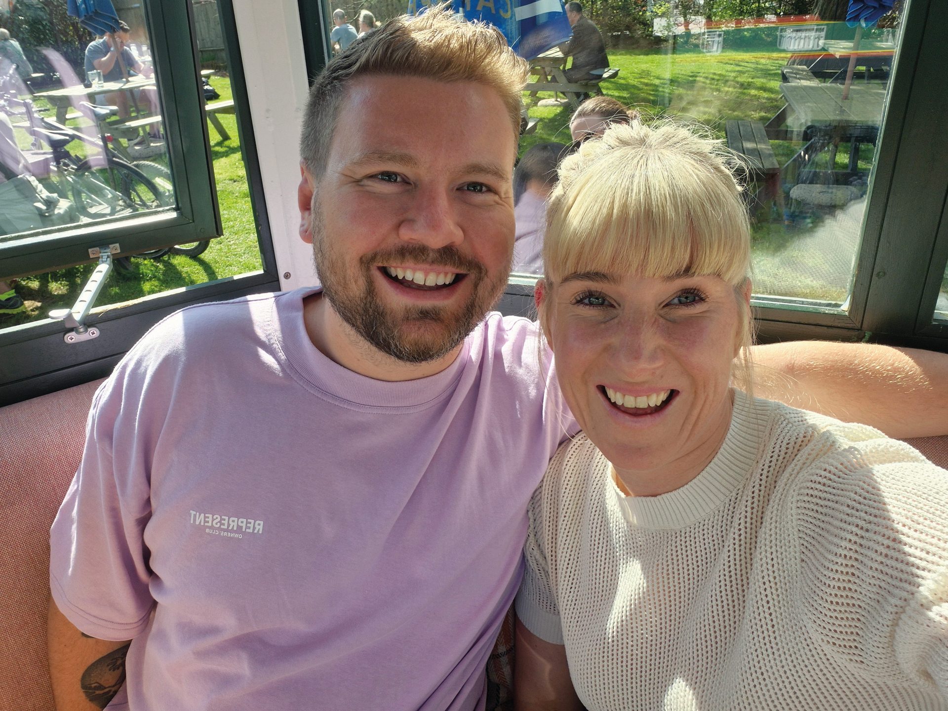 Selfie of a smiling man and woman indoors, with an outdoor garden view through a window.